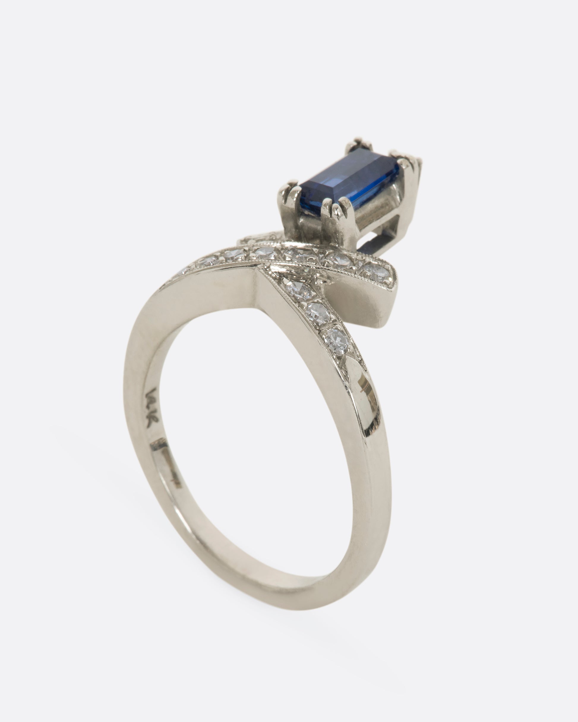 A white gold crossed band ring with a baguette blue sapphire topped with a diamond. View standing from the side.