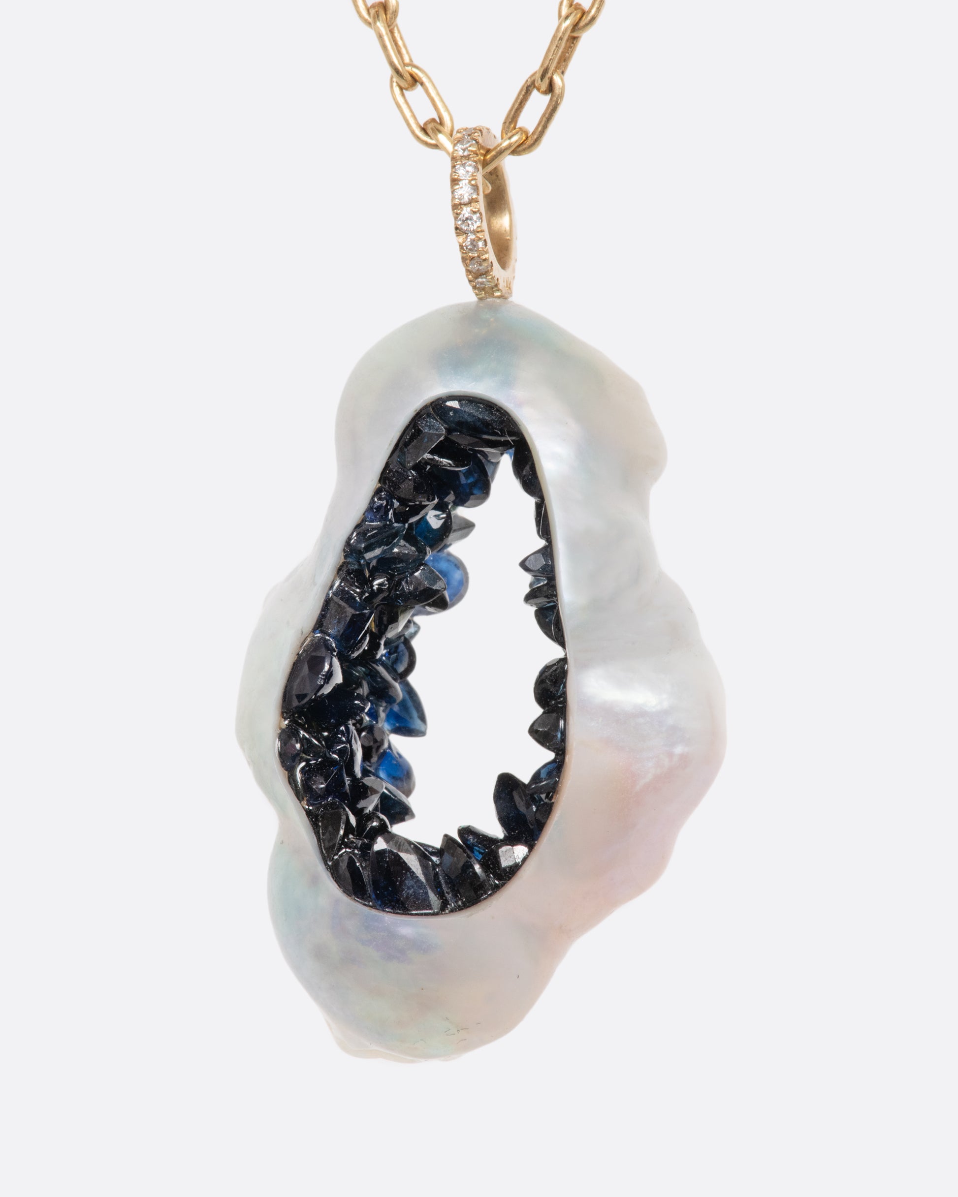 A yellow gold necklace with a hollowed souffle pearl pendant filled with a blue sapphire geode hanging from a pave diamond bale. Shown from the side.