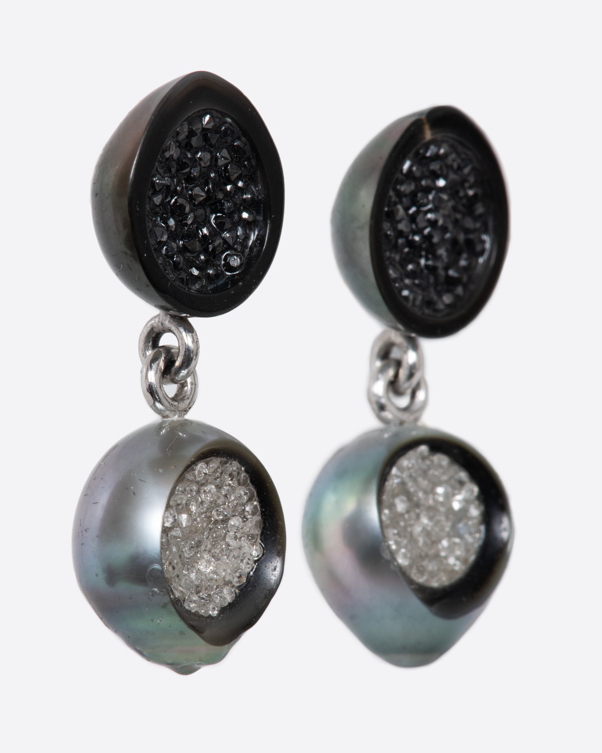 A pair of white gold hollowed out pearl drop earrings with black and white diamonds filling each. Shown from the side.