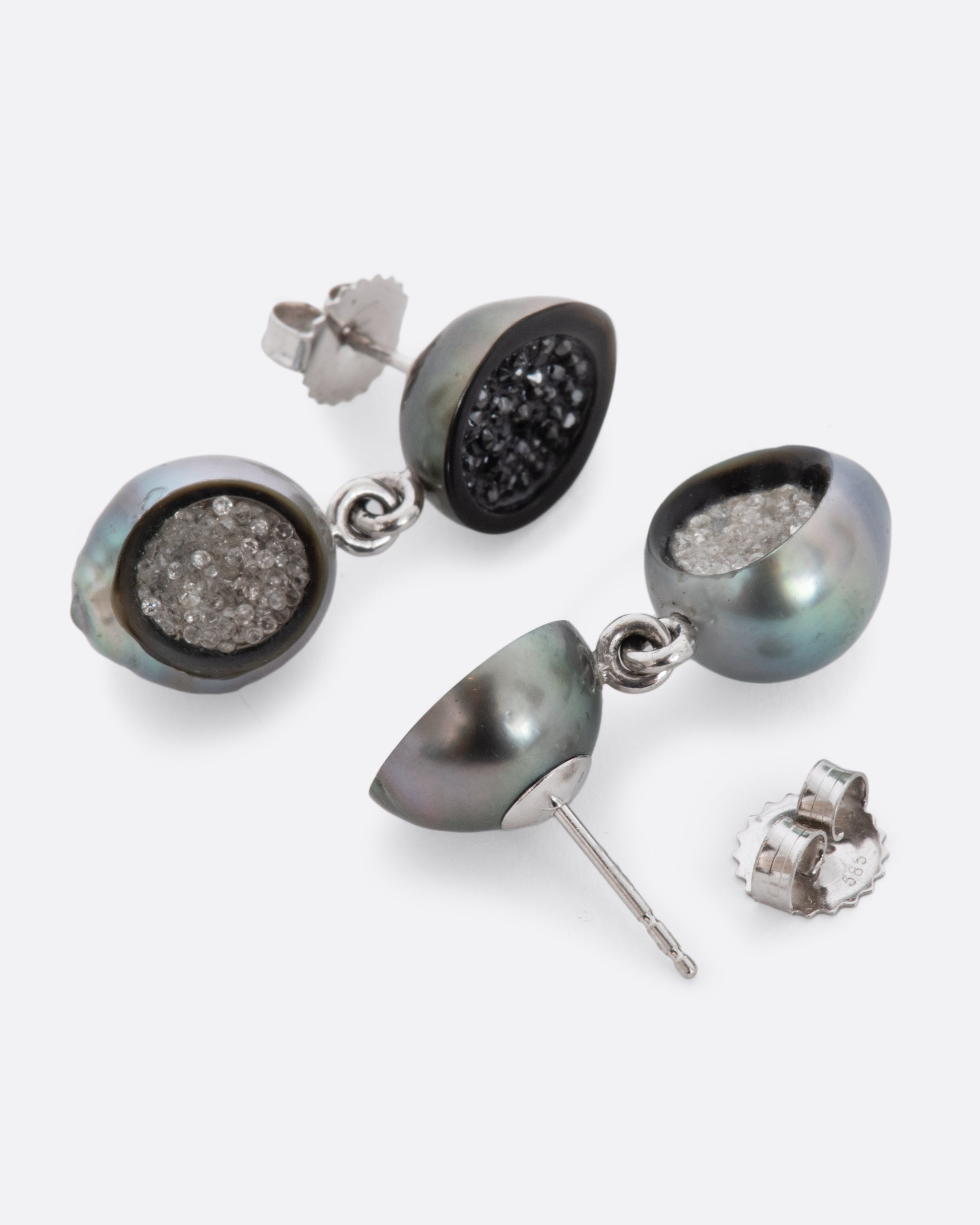 A pair of white gold hollowed out pearl drop earrings with black and white diamonds filling each. Shown laying flat.