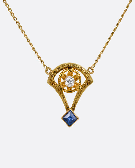 A yellow gold cable chain necklace with a pendant shaped like the basket of a mini hot air balloon with a prong set diamond. and princess cut sapphire. Shown from the front.