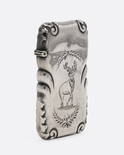 A silver match safe with a deer among scroll work, shown from the side.