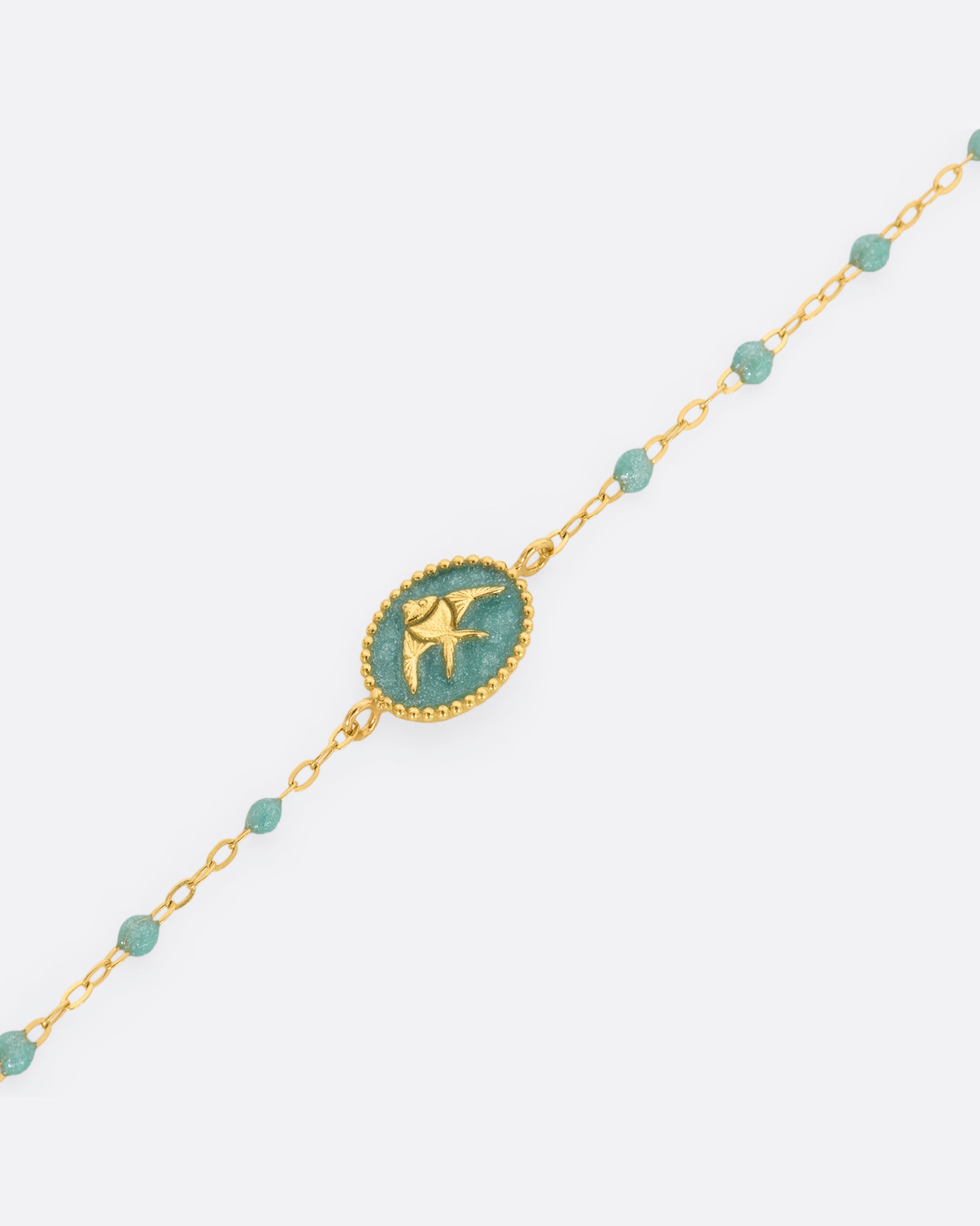 A classic yellow gold Gigi Clozeau bracelet with sparkly iceberg blue resin beads and an oval tag pendant, with a gold angel fish at its center. View of the pendant close up.