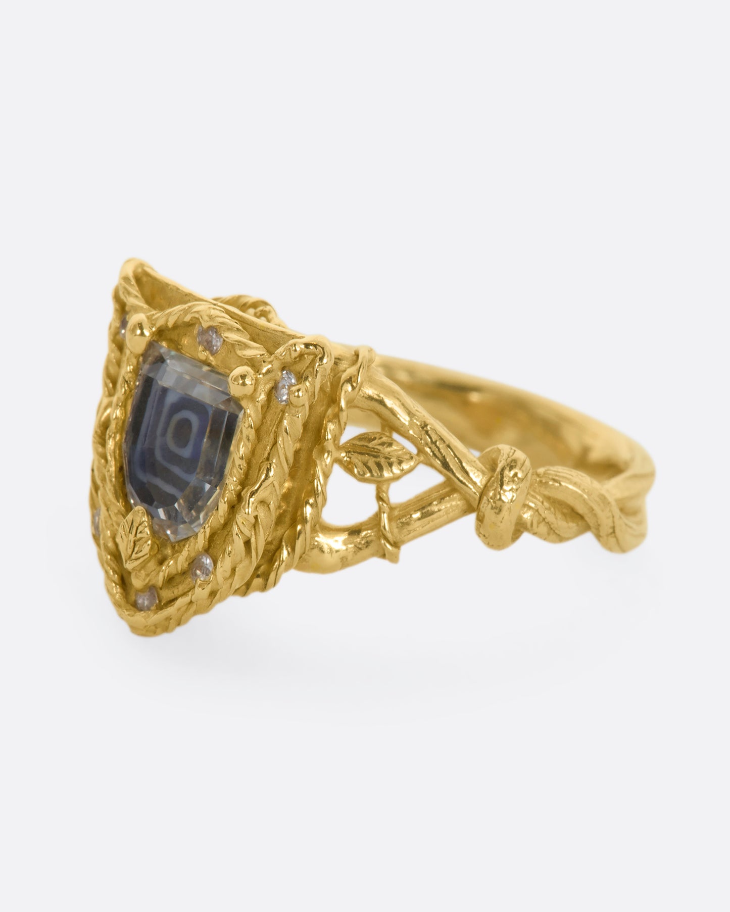 A yellow gold shield shaped ring with vine motif and a geometric portrait cut sapphire over Italian micromosaic glass with brilliant cut white sapphires in a halo around. View from the side.