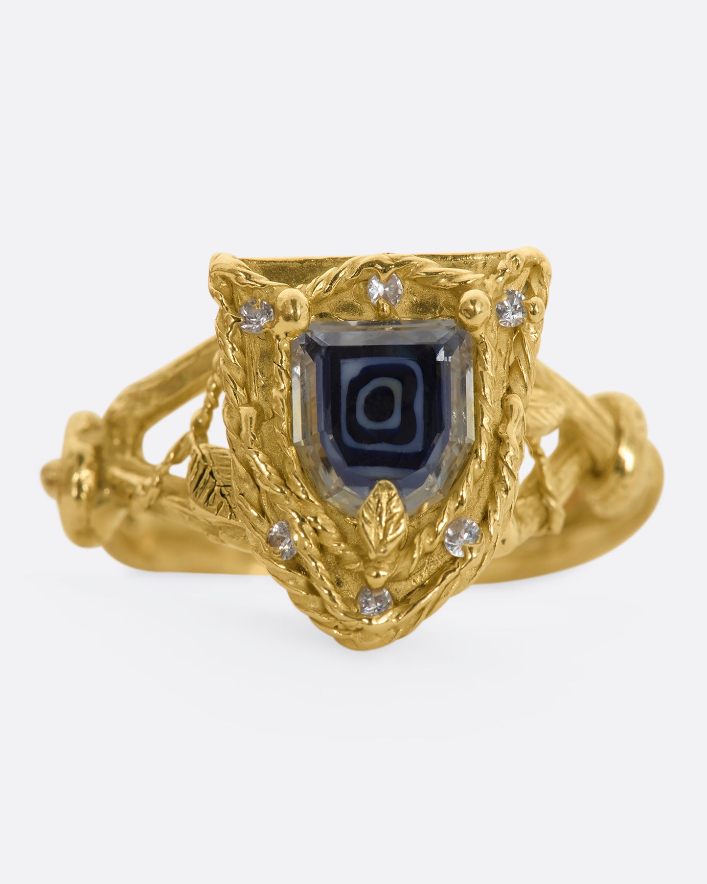 A yellow gold shield shaped ring with vine motif and a geometric portrait cut sapphire over Italian micromosaic glass with brilliant cut white sapphires in a halo around. View close up, from the front.