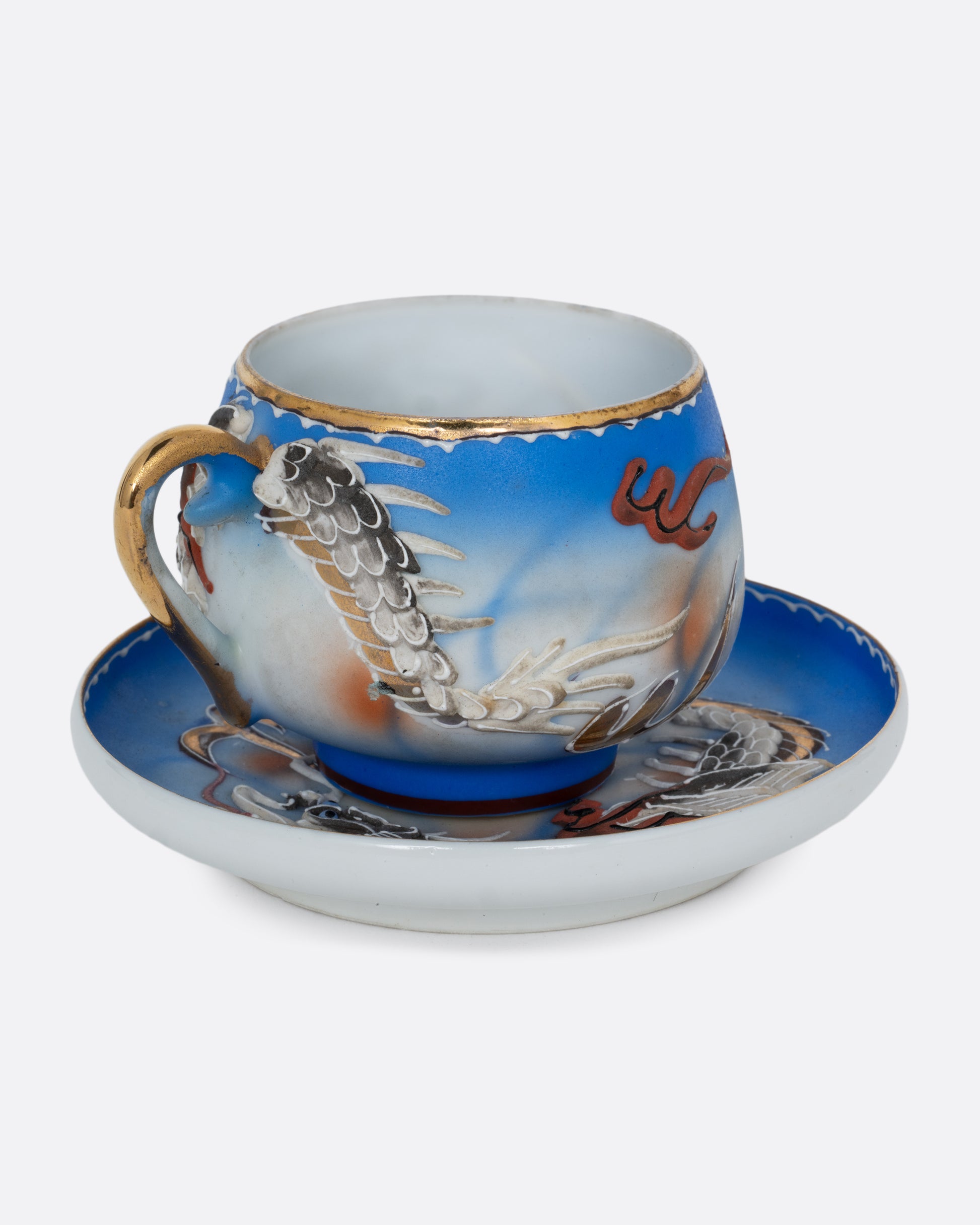 A dragonware moriage teacup and saucer with an enamel dragon motif in vibrant hues. A lithophane nude figure sits at the base of the cup. Shown from the side.