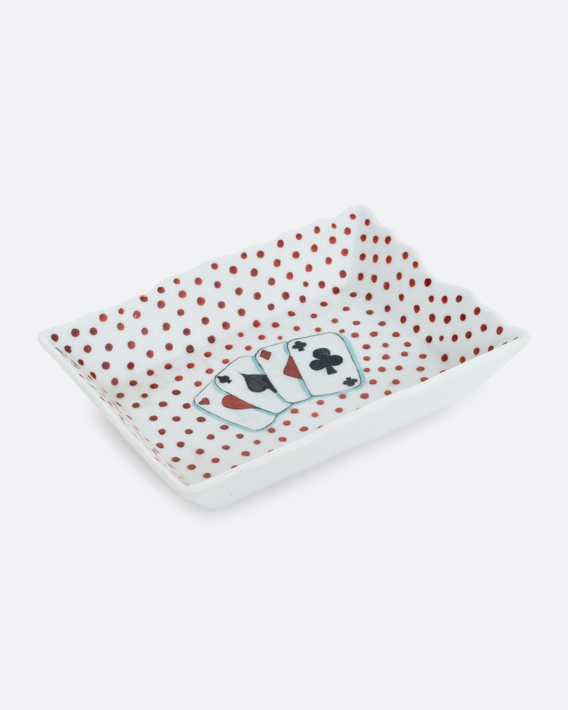 A white rectangular ceramic dish with red hand painted polkadots, and four playing cards at the center. Shown at an angle from the top.