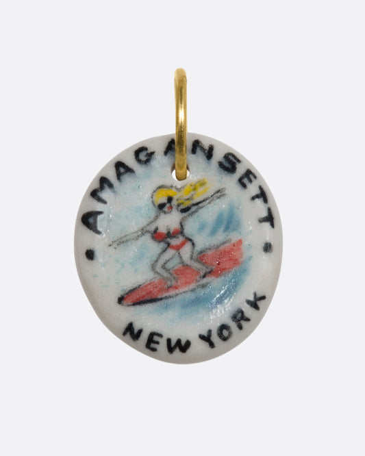 A hand painted porcelain charm with a blond woman in a bikini surfing on it and the words "Amagansett New York". View from the front.