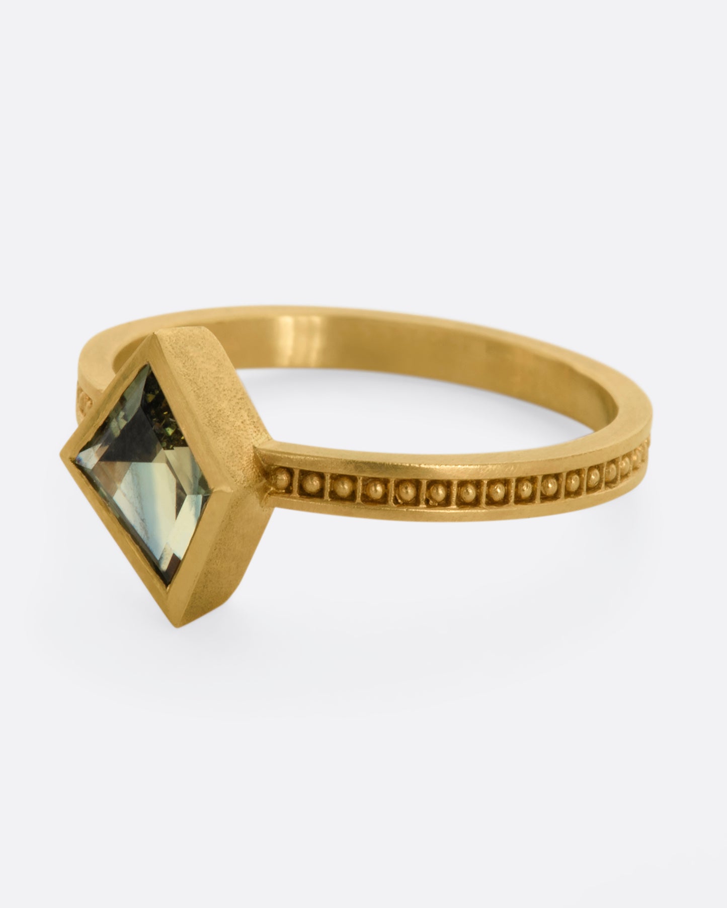 A yellow gold ring with a trapezoid green sapphire and raised dots in square shaped holes around the band. View from the alternate side.