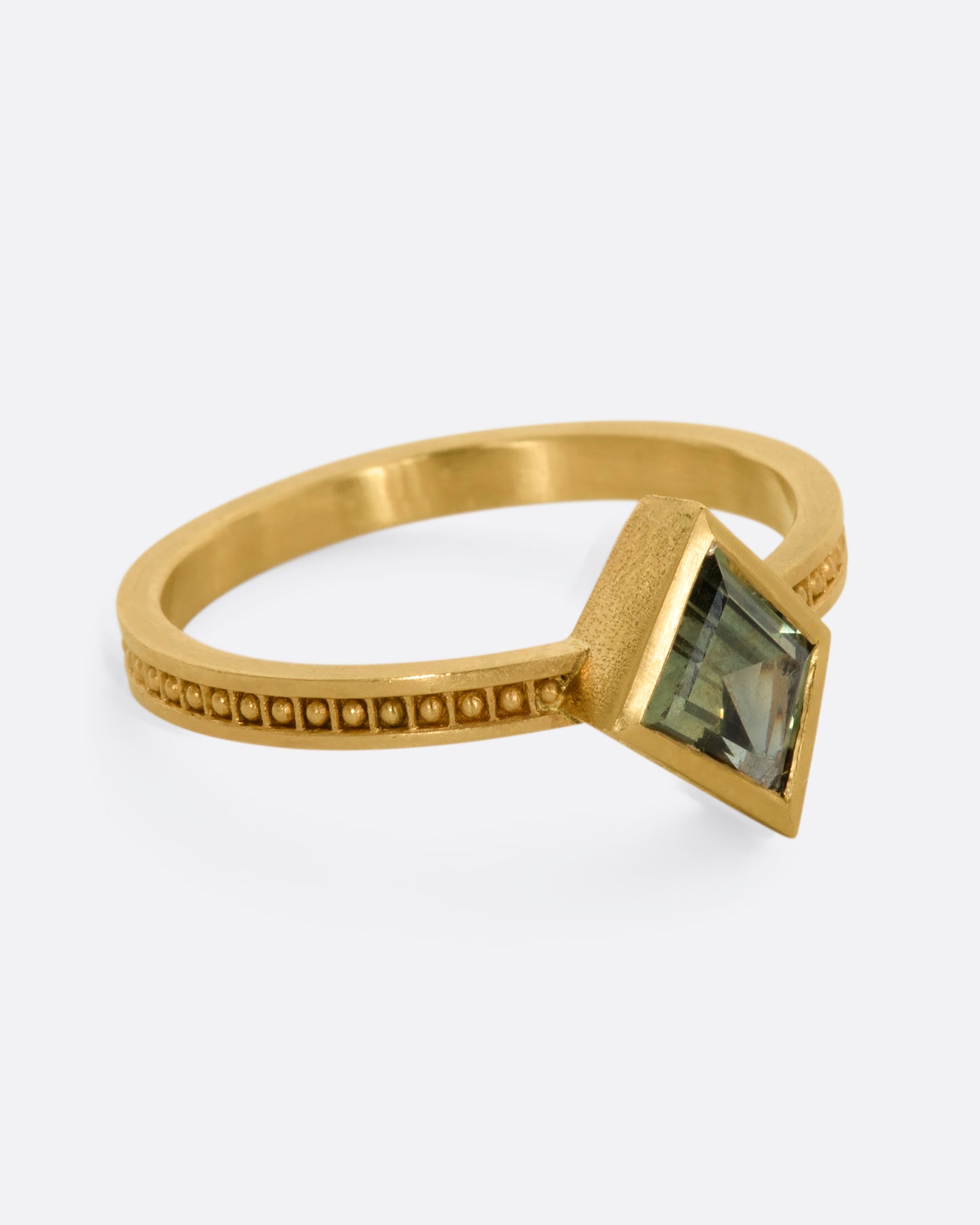 A yellow gold ring with a trapezoid green sapphire and raised dots in square shaped holes around the band. View from the side.