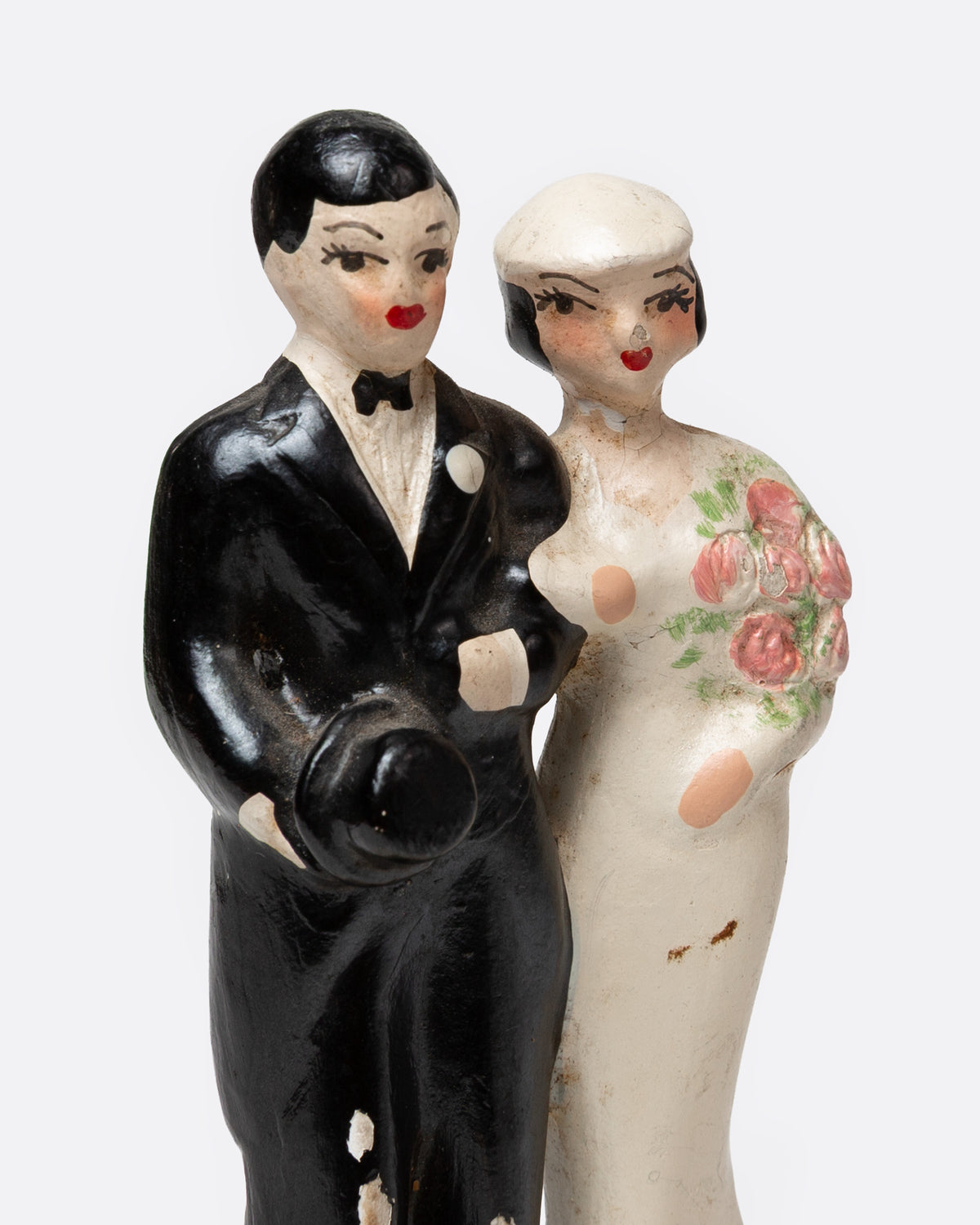 A close up of a ceramic wedding cake topper with a groom in a black tuxedo and a bride in a cream gown with pink floral details, shown from the front.