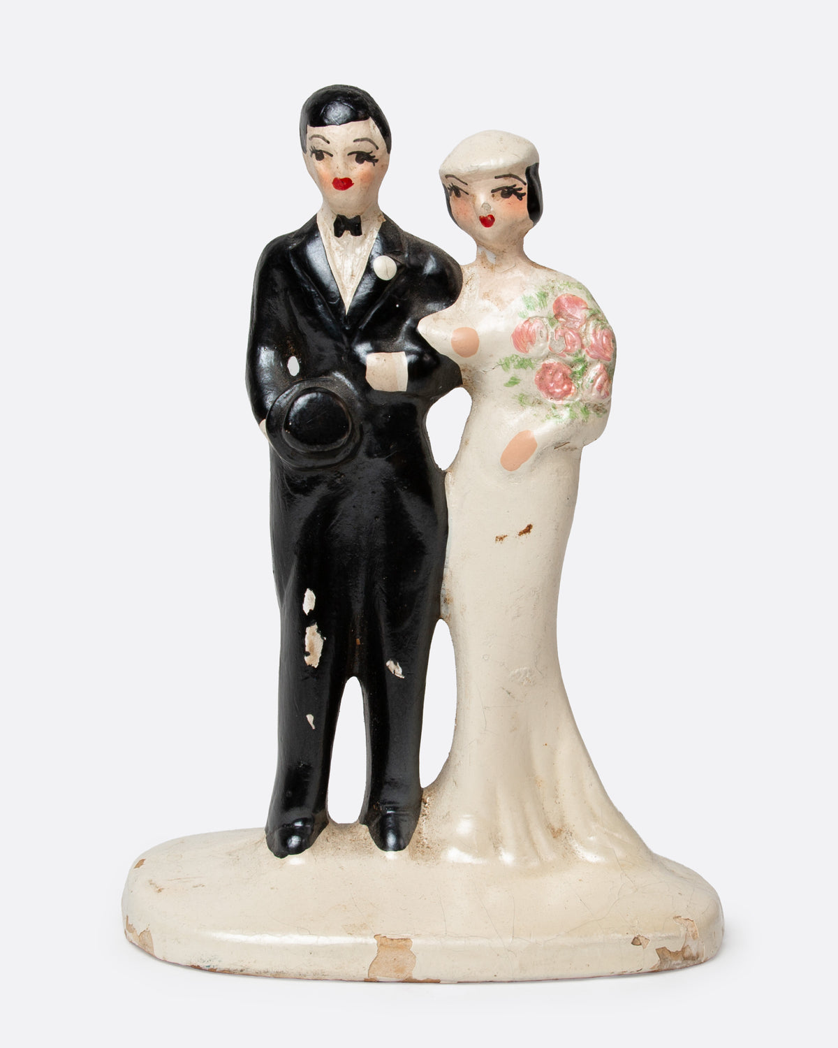 A ceramic wedding cake topper with a groom in a black tuxedo and a bride in a cream gown with pink floral details, shown from the front.