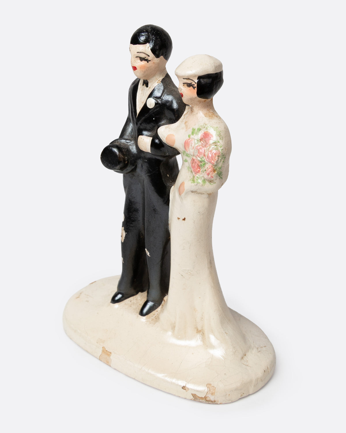 A ceramic wedding cake topper with a groom in a black tuxedo and a bride in a cream gown with pink floral details, shown from the side.