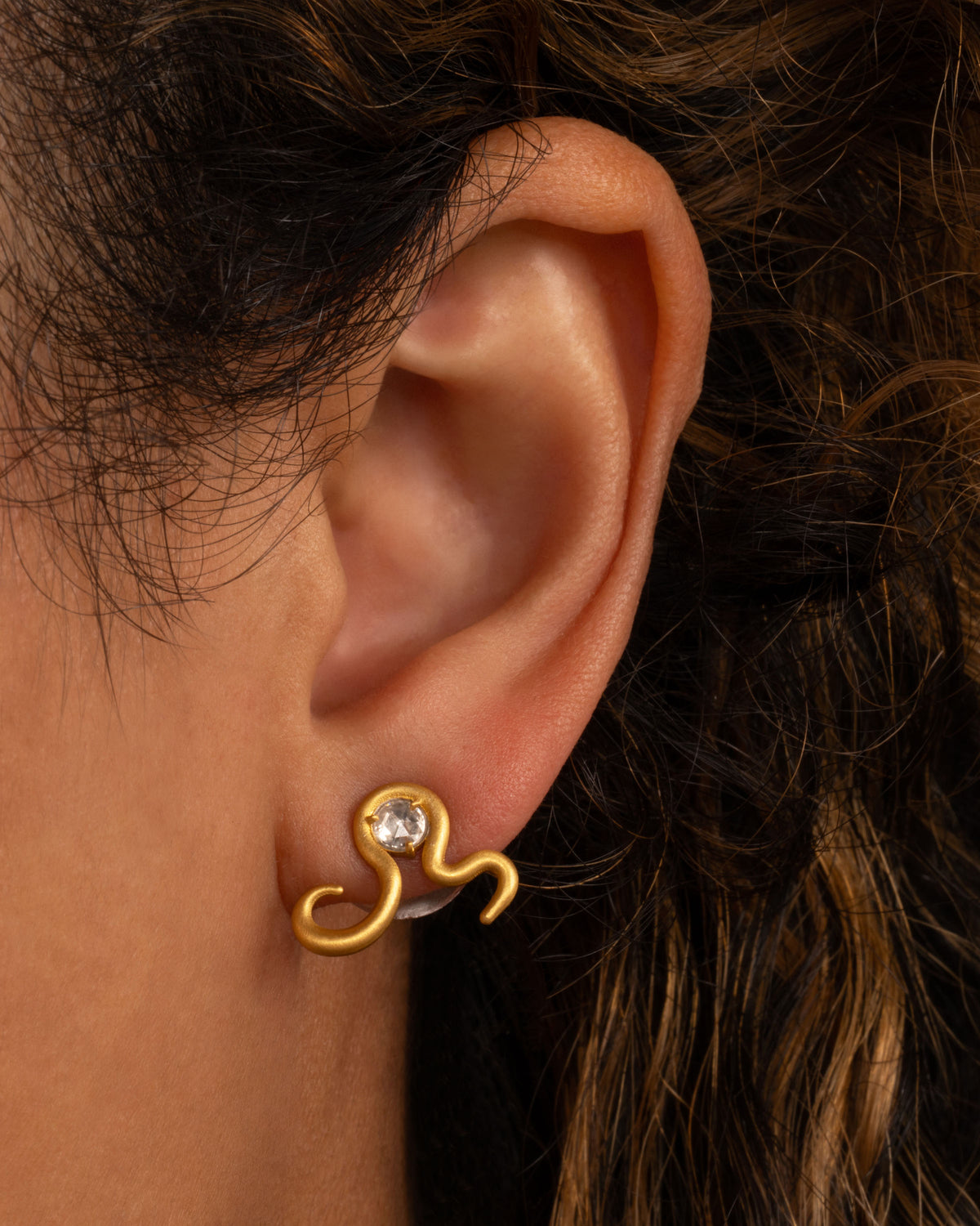 Lori Leven snake stud earring in matte yellow gold worn on an ear, a curving gold snake shape set with a rose-cut diamond