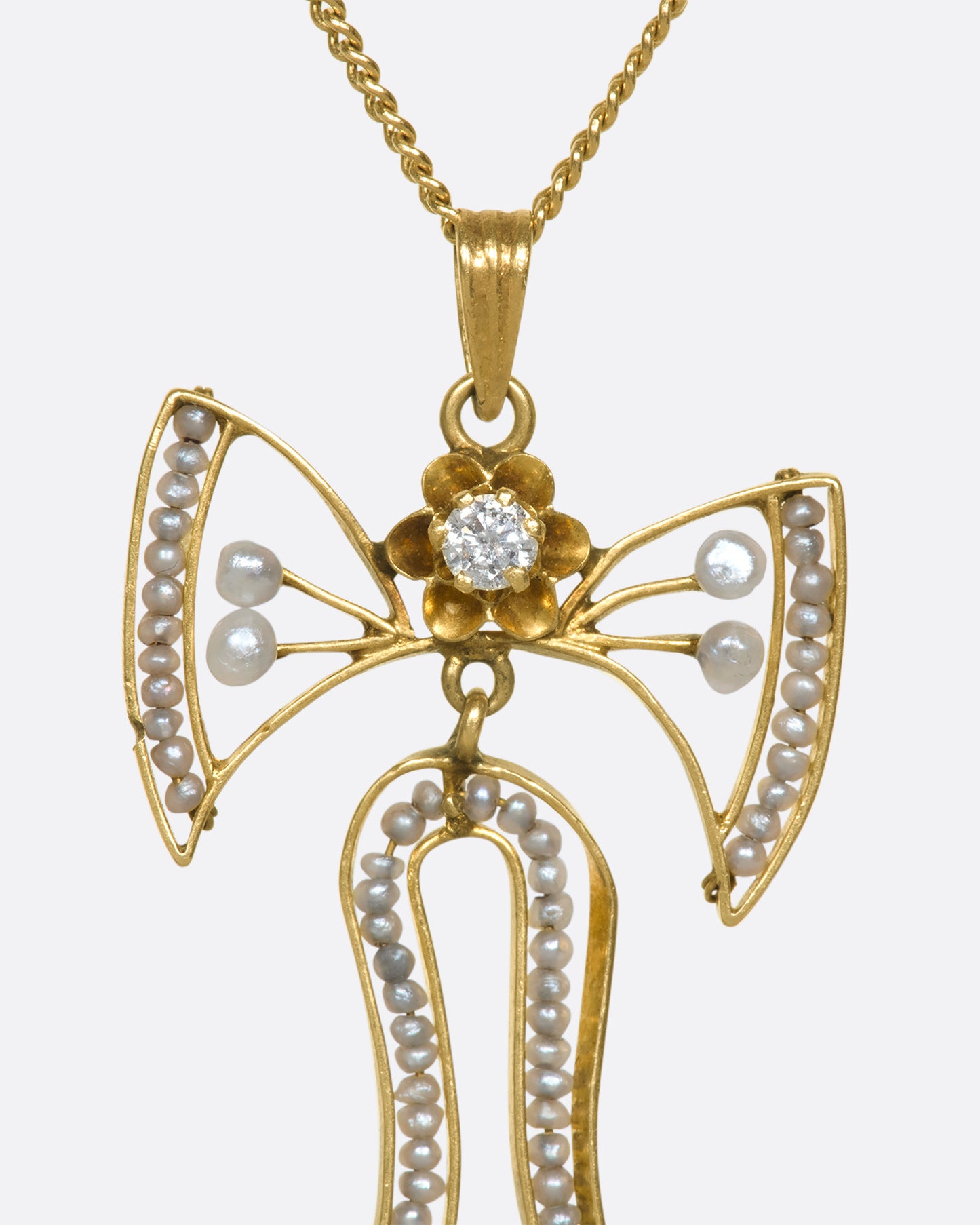 A close up view of a the top half of a yellow gold bow pendant with seed pearls and a diamond.