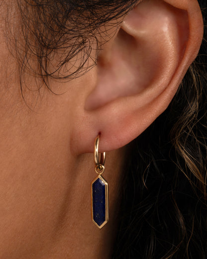 A yellow gold hoop earring with a long hexagonal lapis dangle, shown on an ear.