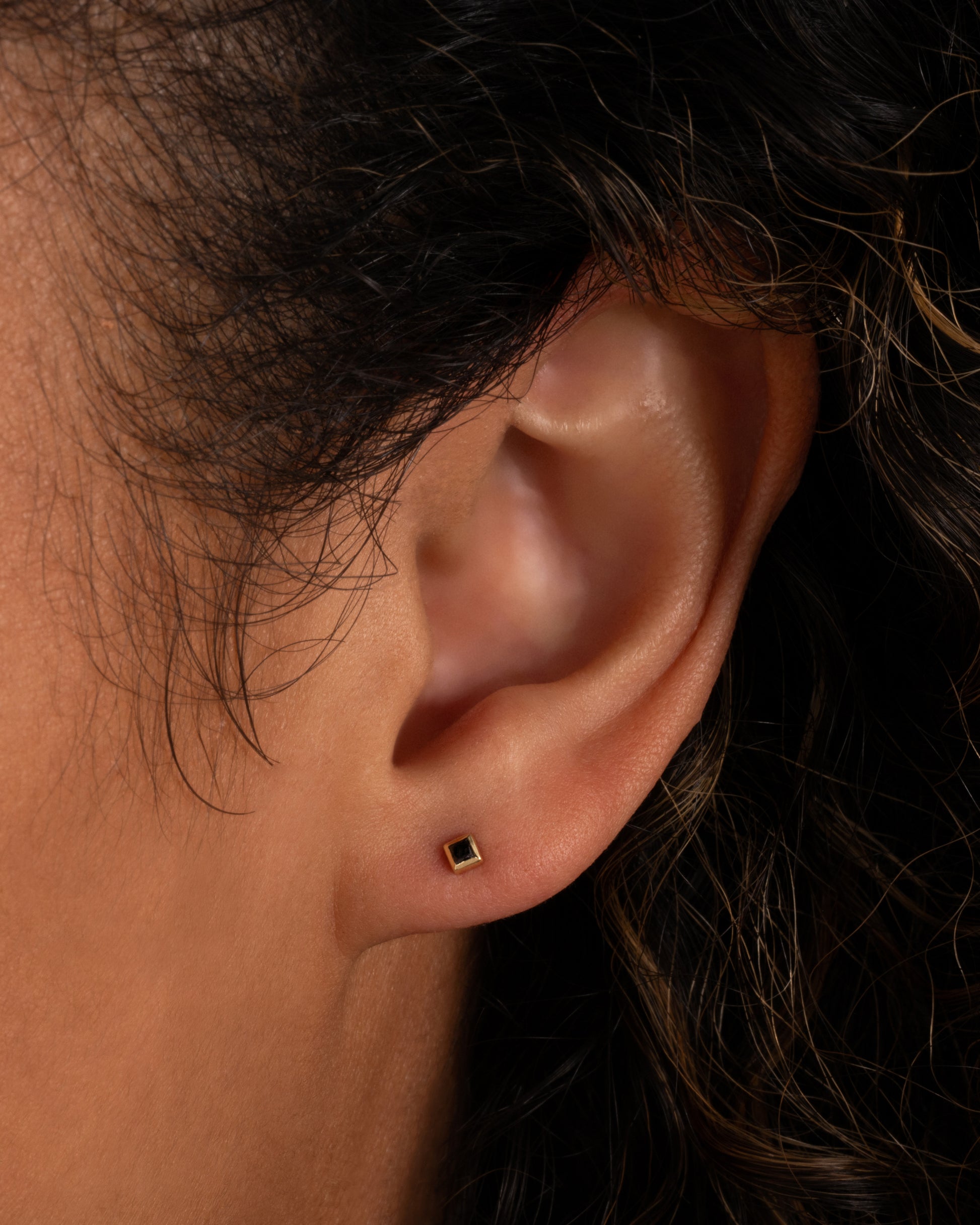 A yellow gold stud earring with a princess cut black diamond. Shown in an ear.