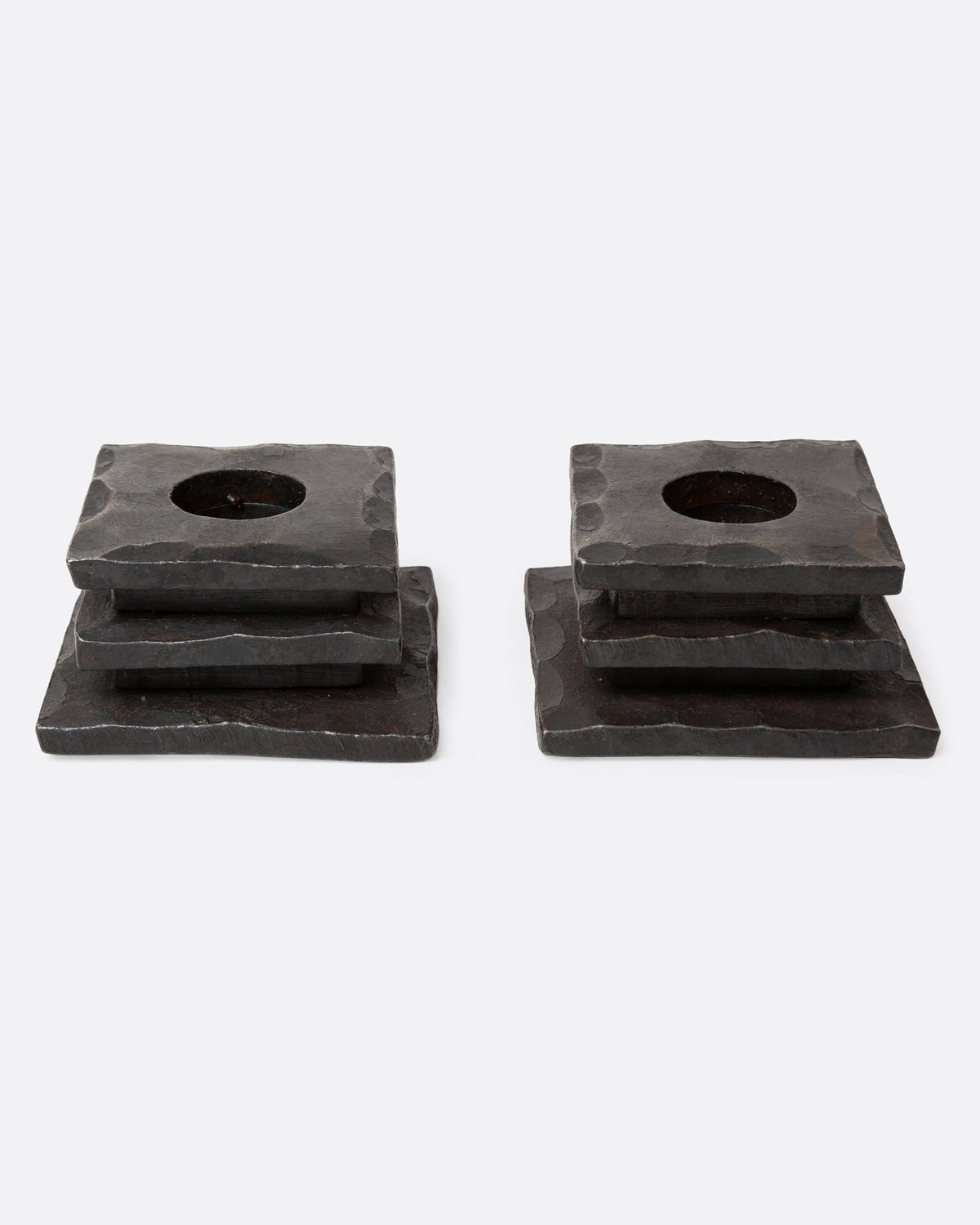 A pair of iron stacked square candle holders with a hammered texture and a circular cutout in the top surface, shown from the front.