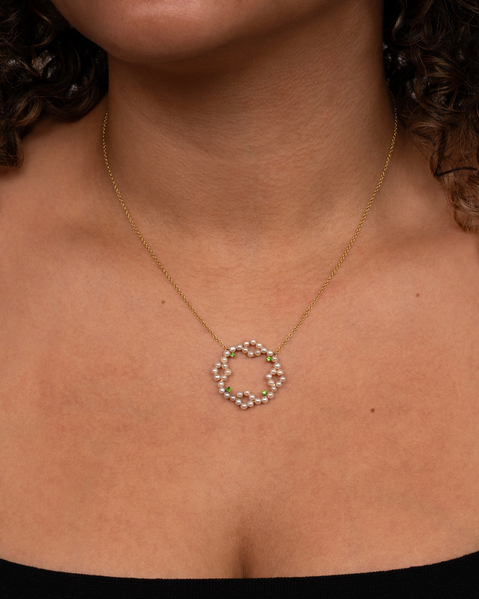 A yellow gold necklace with a pearl and green garnet wreath pendant. Shown worn around a neck.