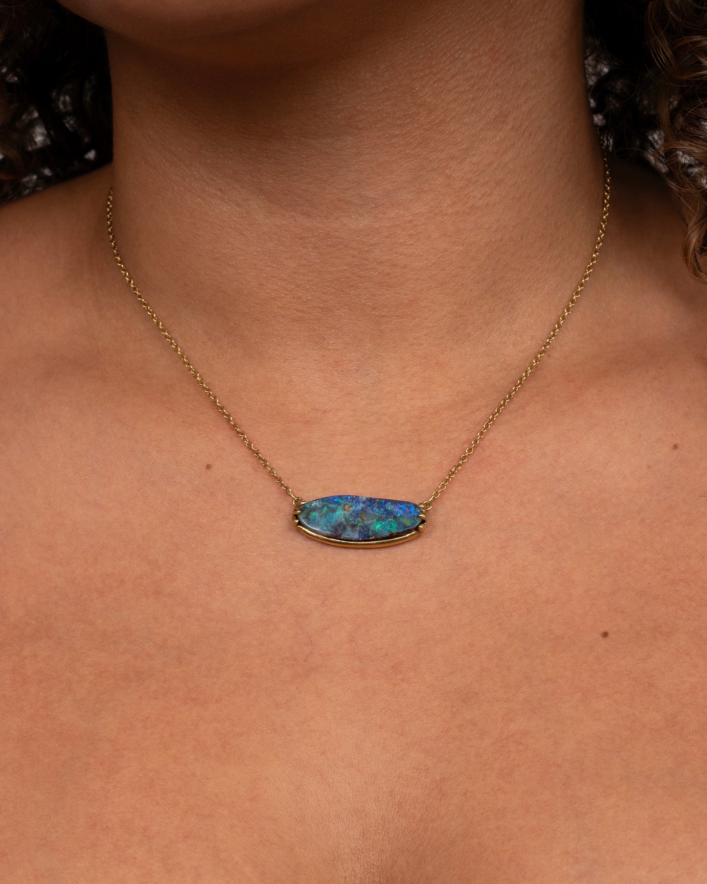 A blue Australian opal sits in a yellow gold east west claw setting and hangs from a yellow gold chain. Shown worn around a neck.