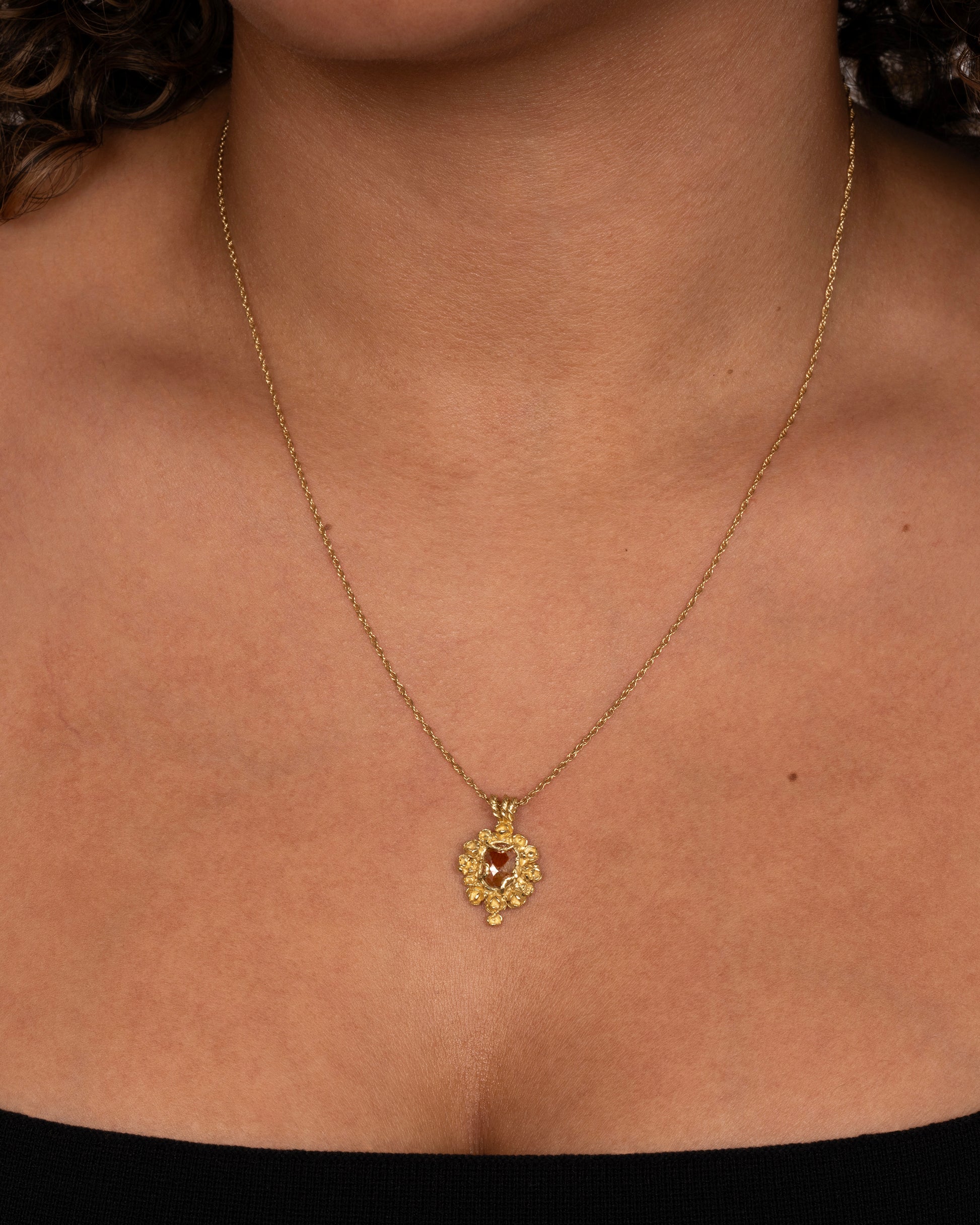 A yellow gold pendant reminiscent of an antique mirror with floral and vine details around an orange rose cut diamond. Shown worn around a neck.