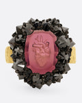A yellow gold ring with a hand carved anatomical heart in a garnet and a dimensional wreath of reverse set black diamonds, shown from the front.
