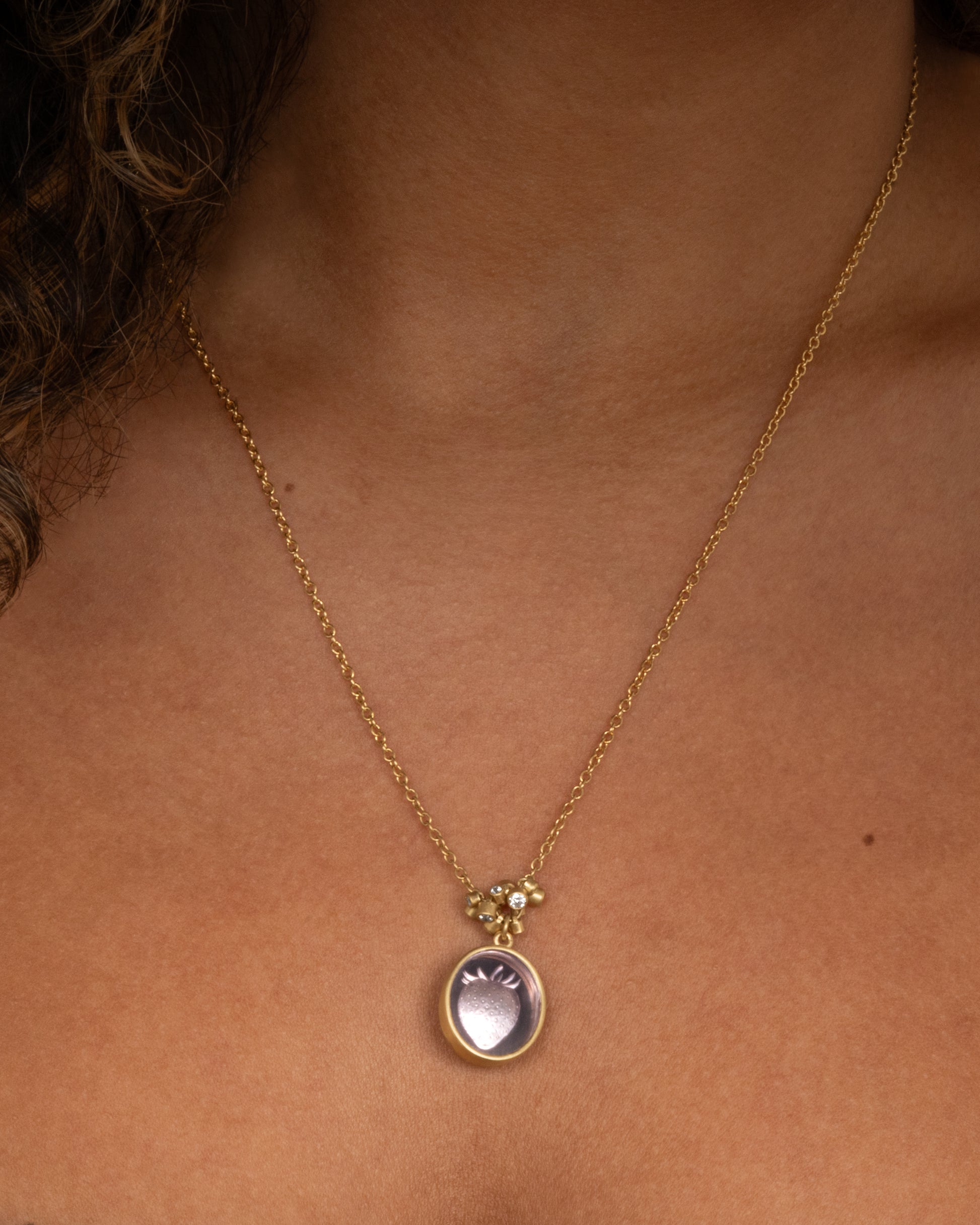 A yellow gold pendant necklace with a amethyst with a hand carved strawberry and a cluster of diamond charms, shown on a person.