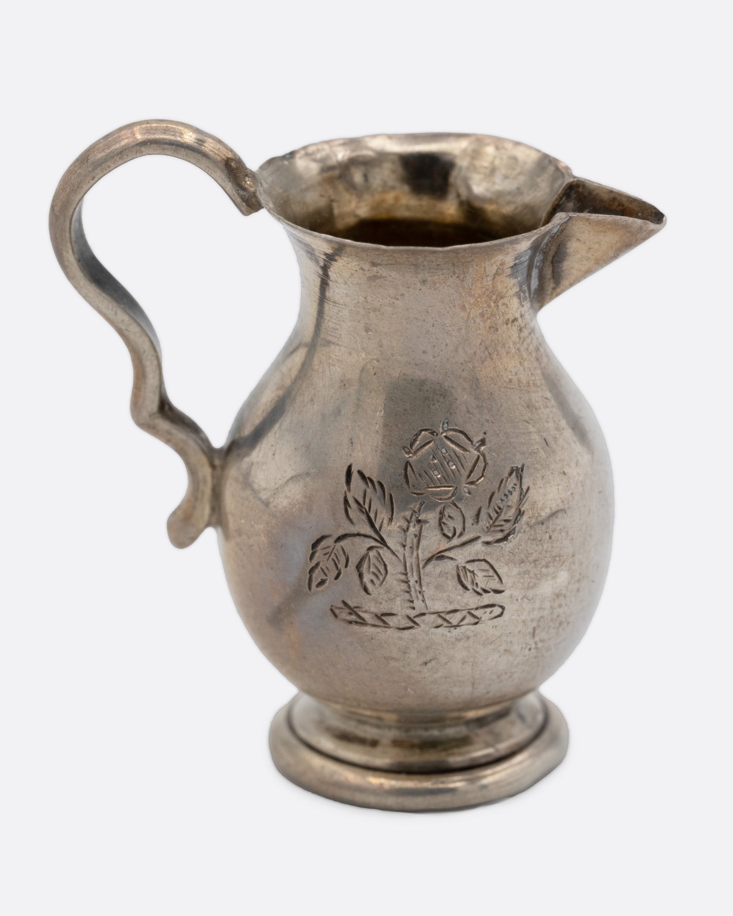 A tiny silver pitcher with an engraved flower, a spout, and handle. Shown from the side.