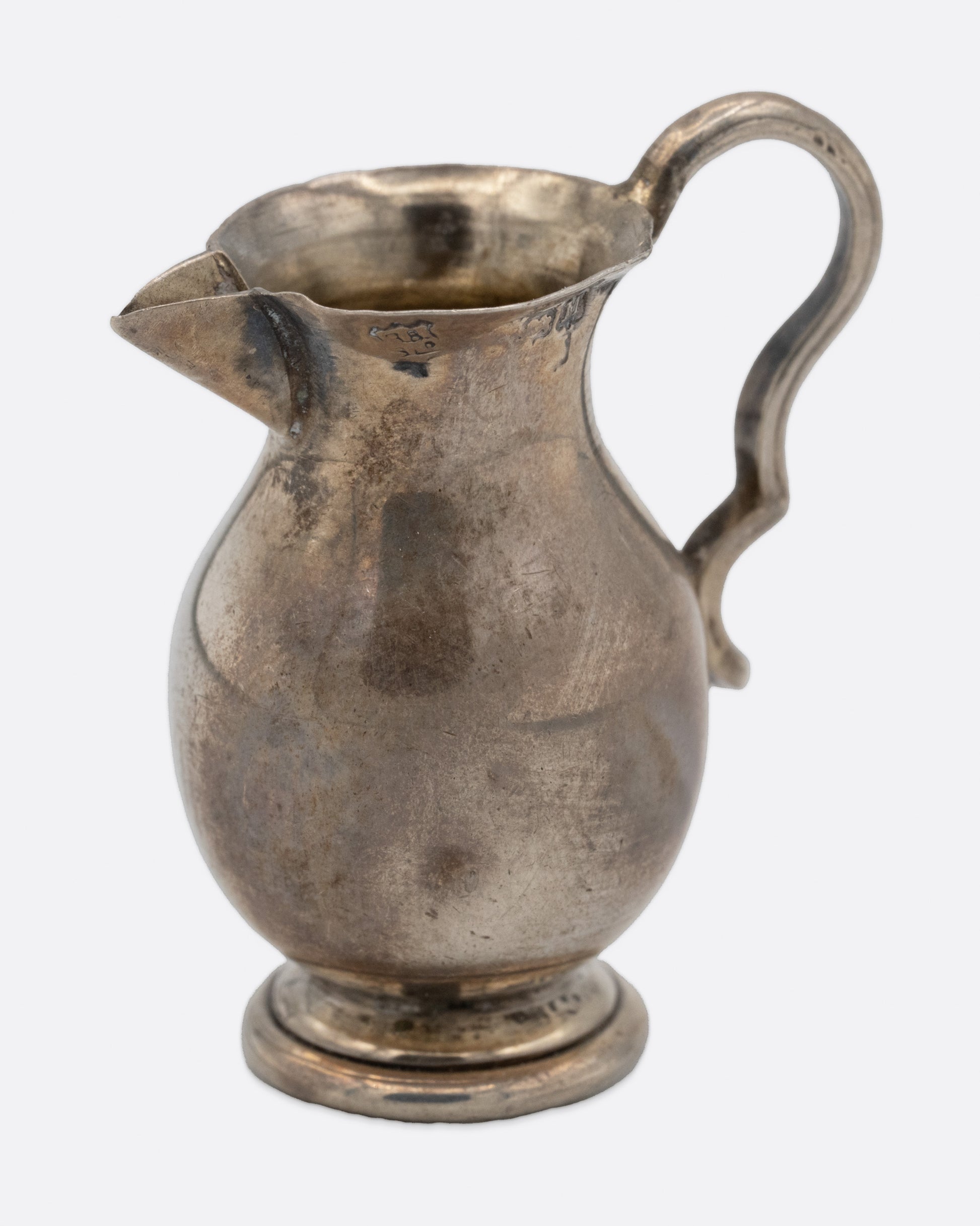 A tiny silver pitcher with an engraved flower, a spout, and handle. Shown from the side.