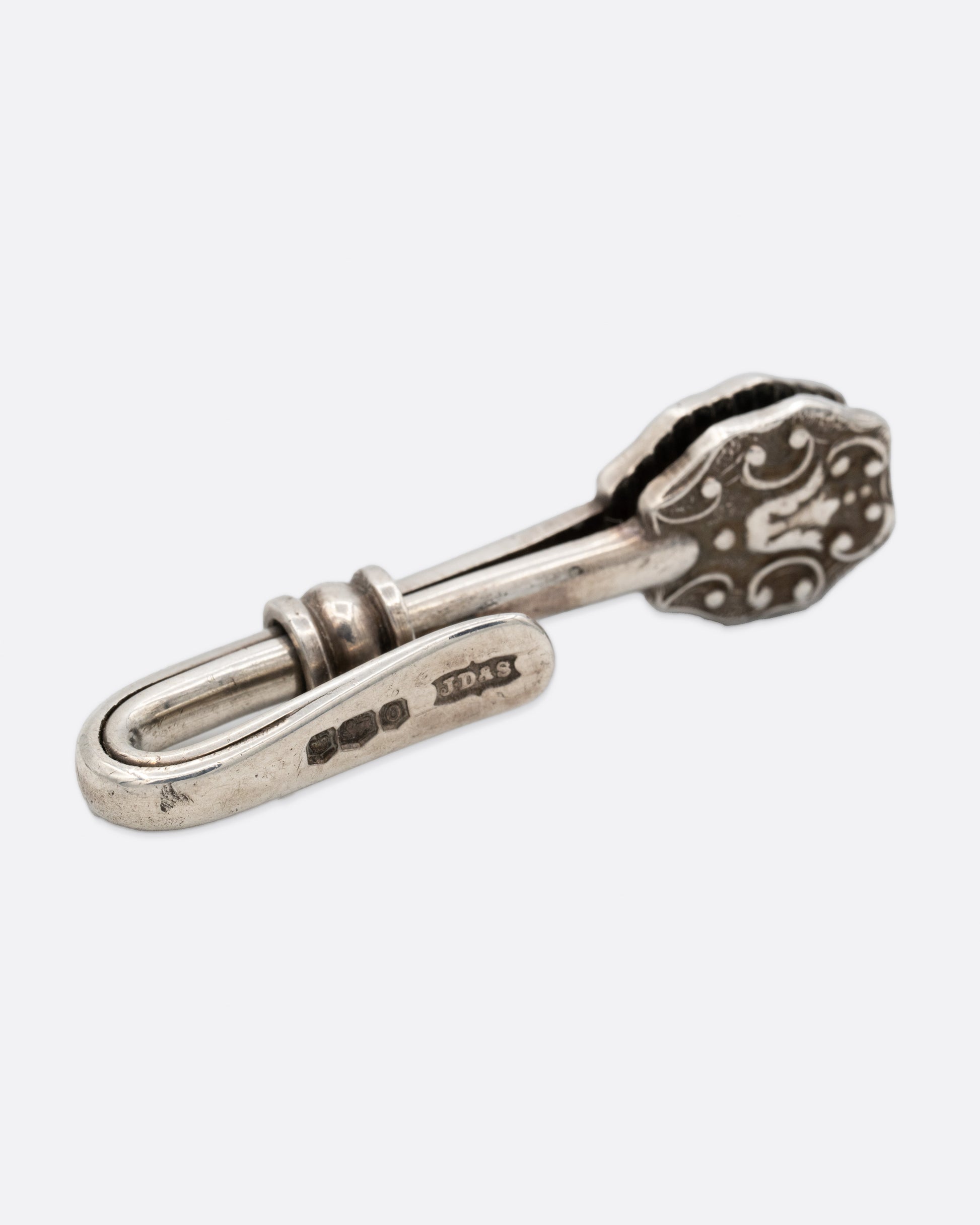 A silver engraved napkin holder with a  hook at one end and an oval clip to hold a napkin at the other. Shown from the back.