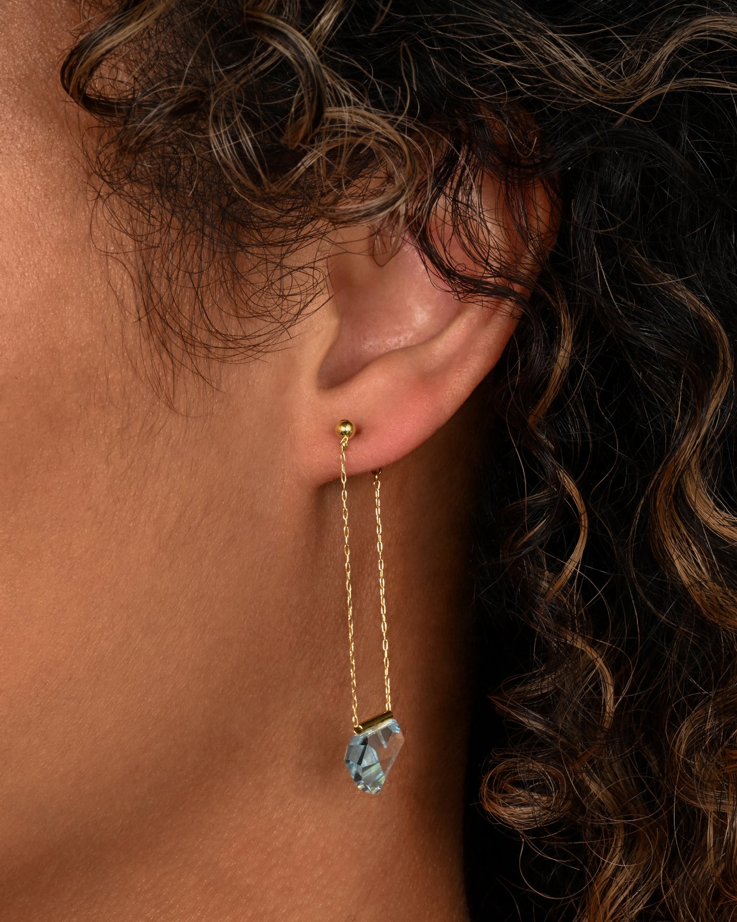 A pair of yellow gold chain earrings with free form blue topaz. Shown on an ear.