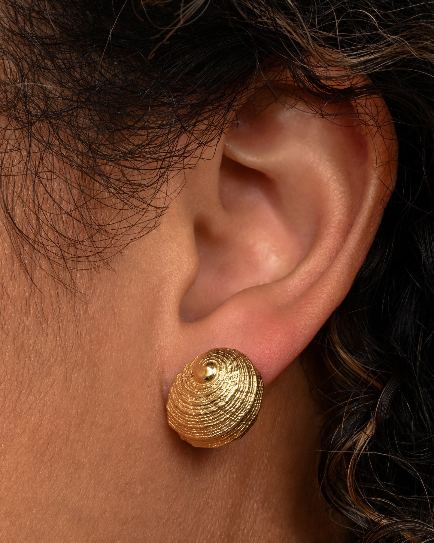 A pair of yellow gold earrings in the shape of a shell. Shown on an ear.