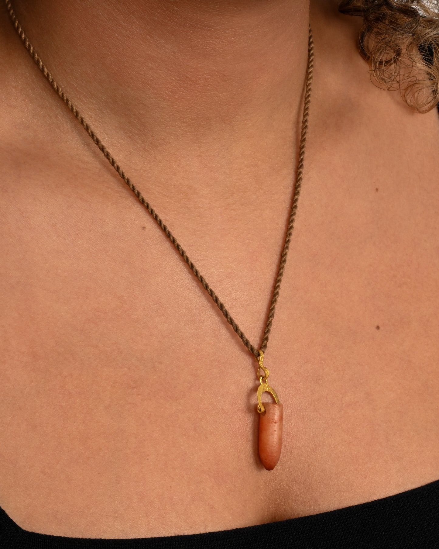 A stunning pre-Columbian carnelian bullet pendant suspended in 22k gold from a lengthy, twisted brown chord. Shown on a neck.