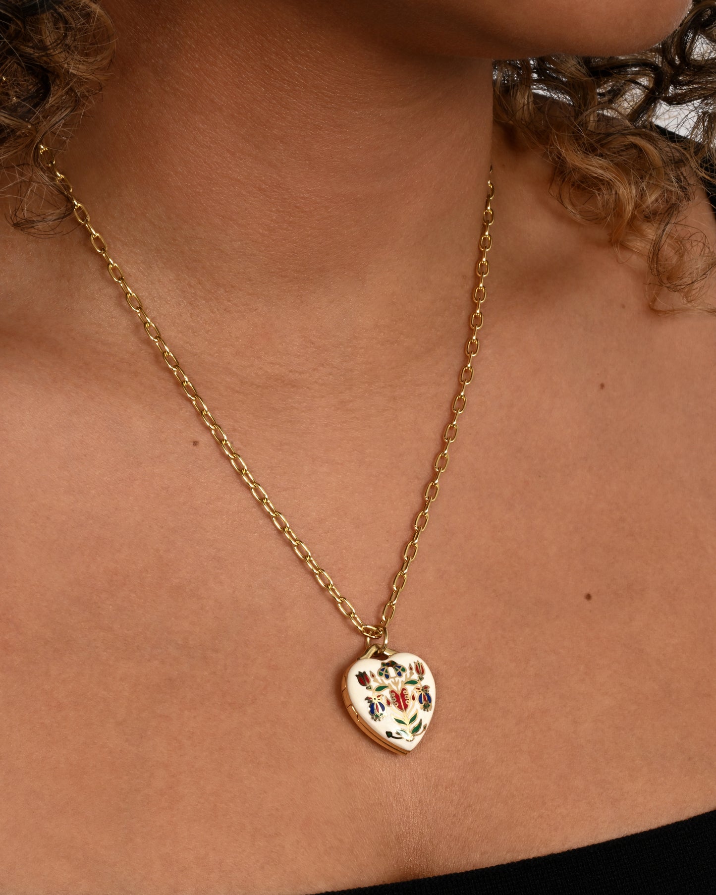 A yellow gold heart shaped locket with a multicolor floral motif on a heavy chain. Shown worn on a neck.