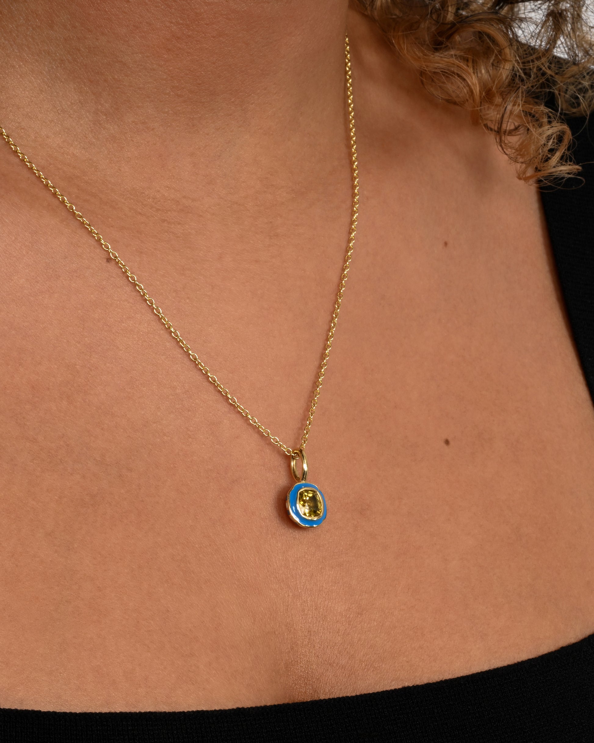 A yellow gold pendant with blue enamel surrounding a cushion cut yellow sapphire. Shown worn on a cable chain.