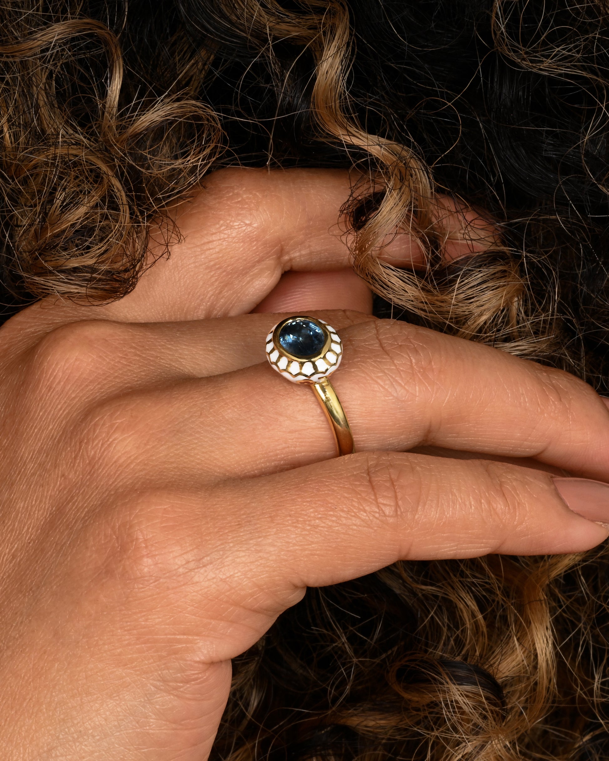 A yellow gold ring with a light blue sapphire and white enamel halo. Shown on a hand.