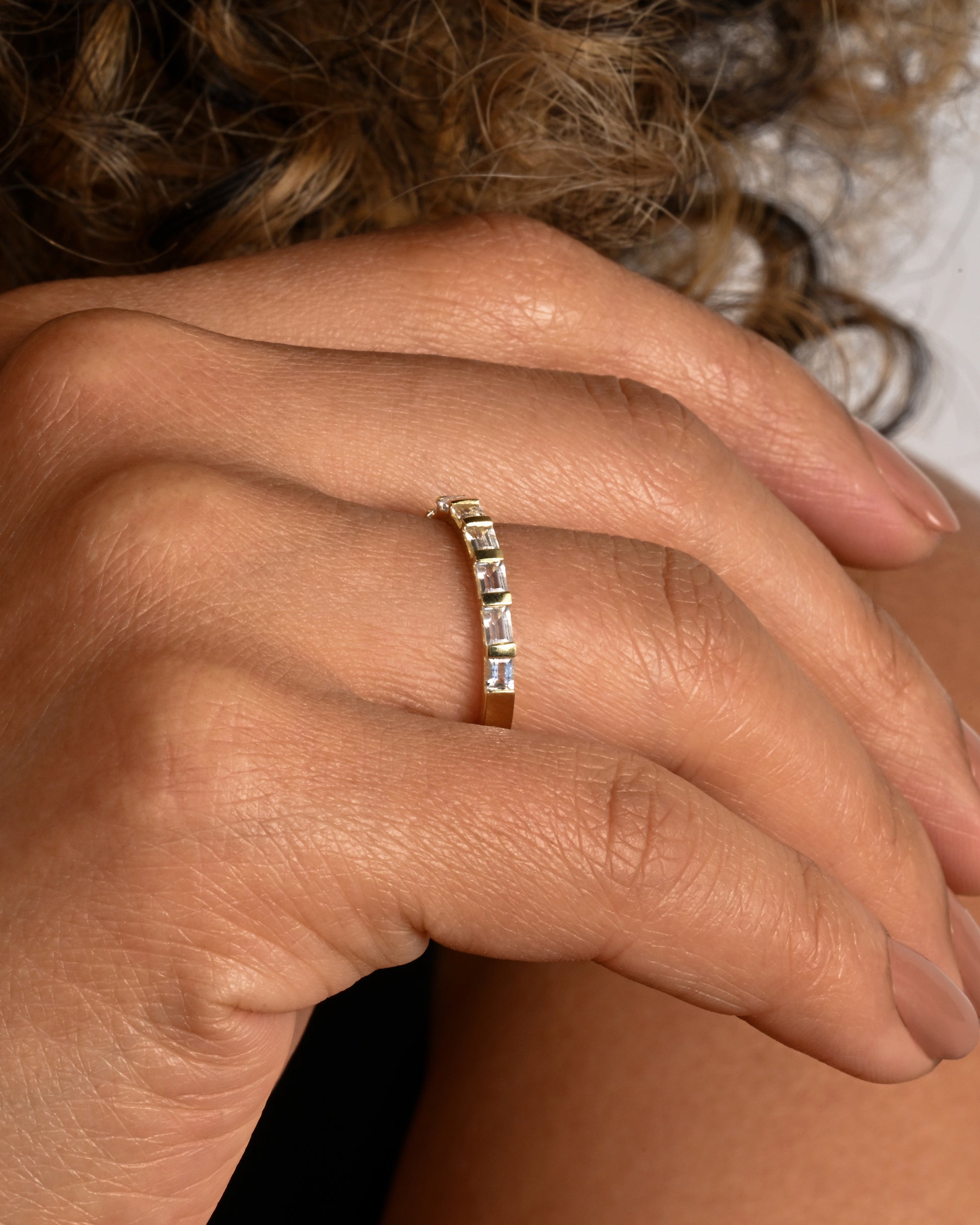 A yellow gold band with seven white sapphire baguettes covering half of the ring. Shown on a hand.