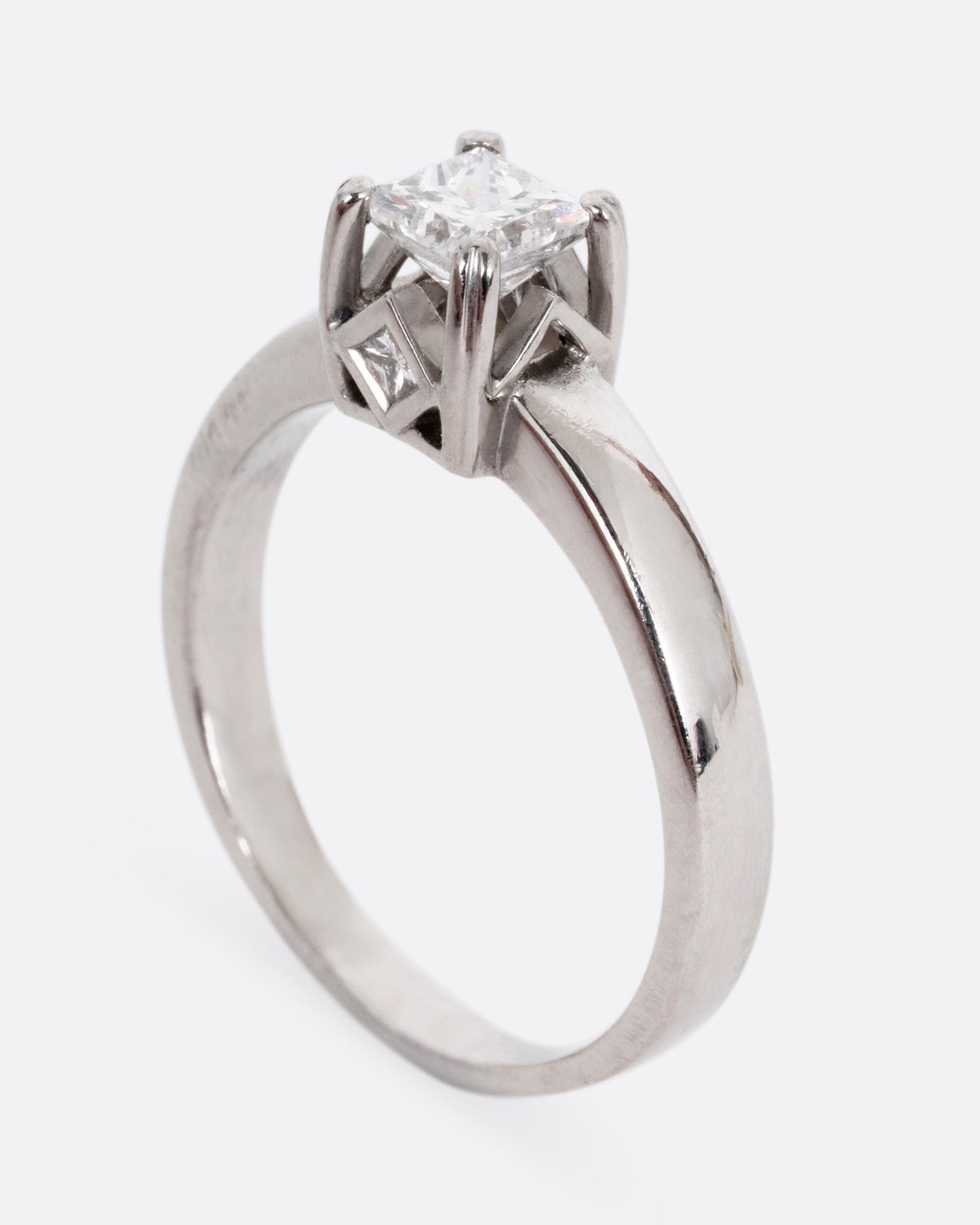 A platinum ring with a princess cut diamond, prong set high above angular cutouts and diamond accents. Shown standing up from the side.