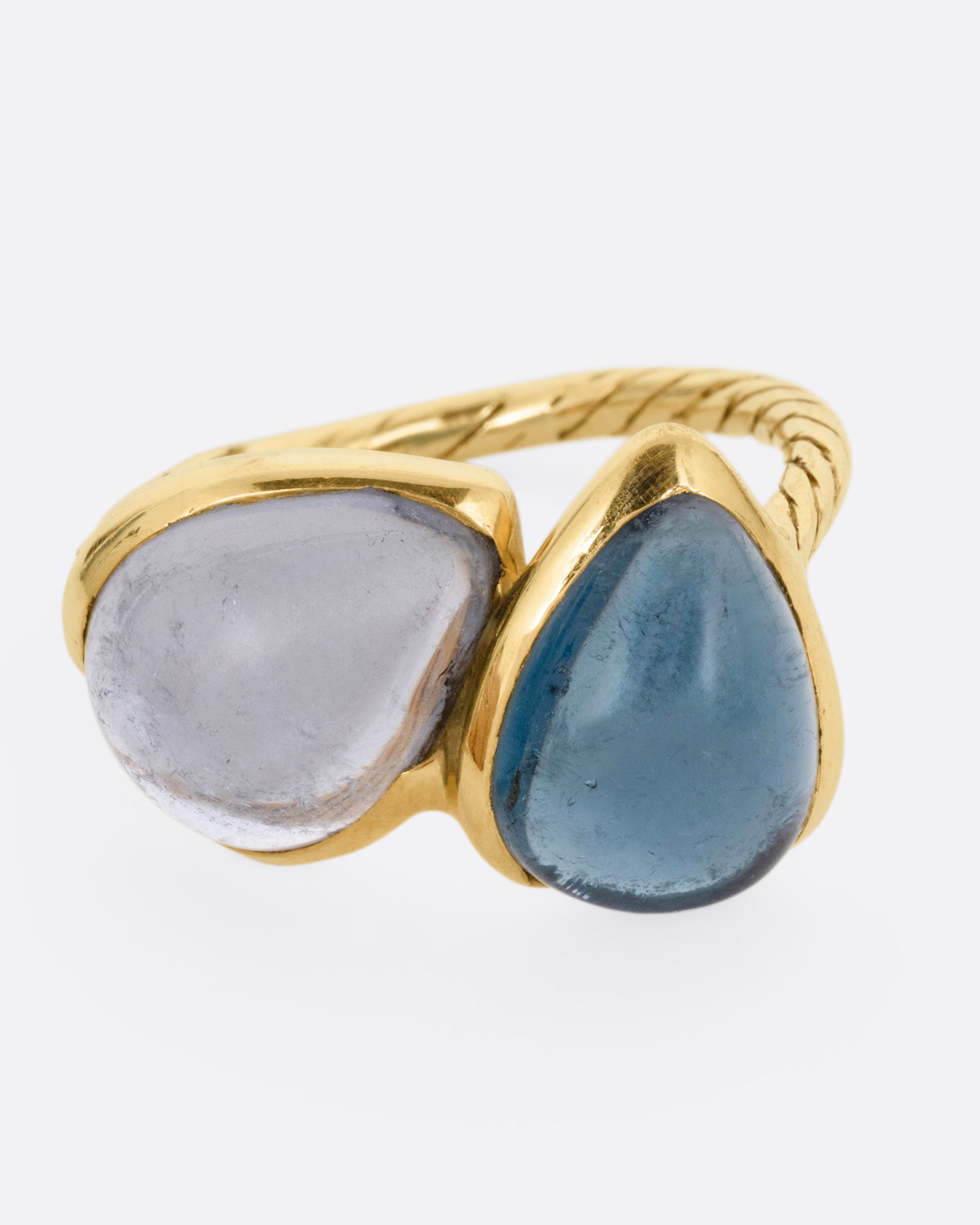 A yellow gold ring with a pair of blue green tourmalines and a textured hand- carved band, shown from the front.
