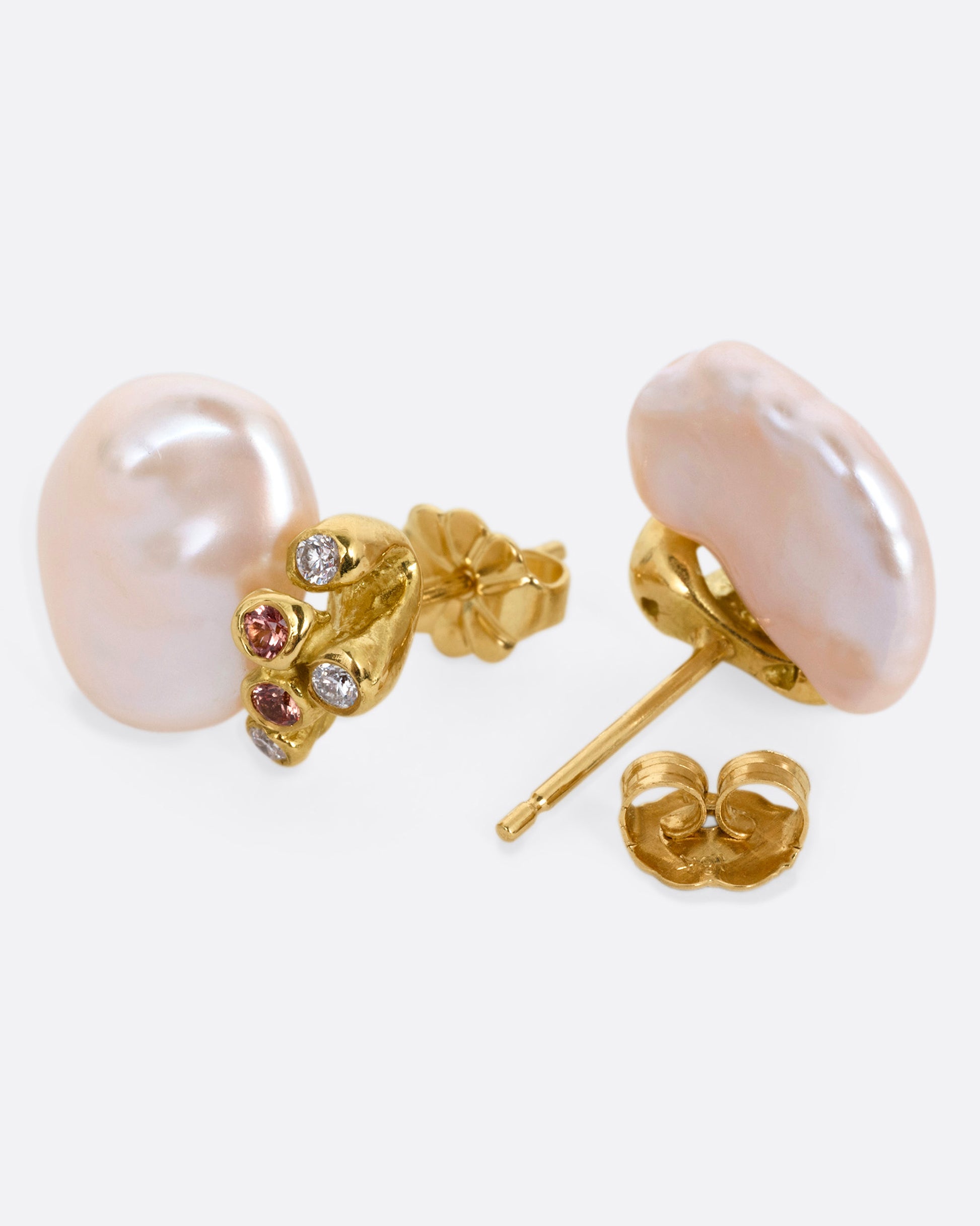 A pair of yellow gold stud earrings with diamond and orange sapphire clusters atop two pink freeform Keshi pearls, shown lying on their sides with one earring open.