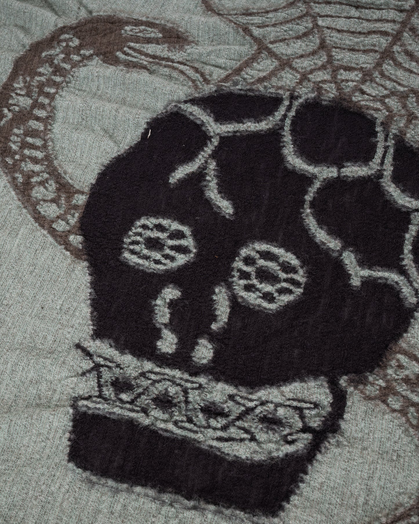 A grey and green wool scarf with a black skull in the center. Close up of the skull.