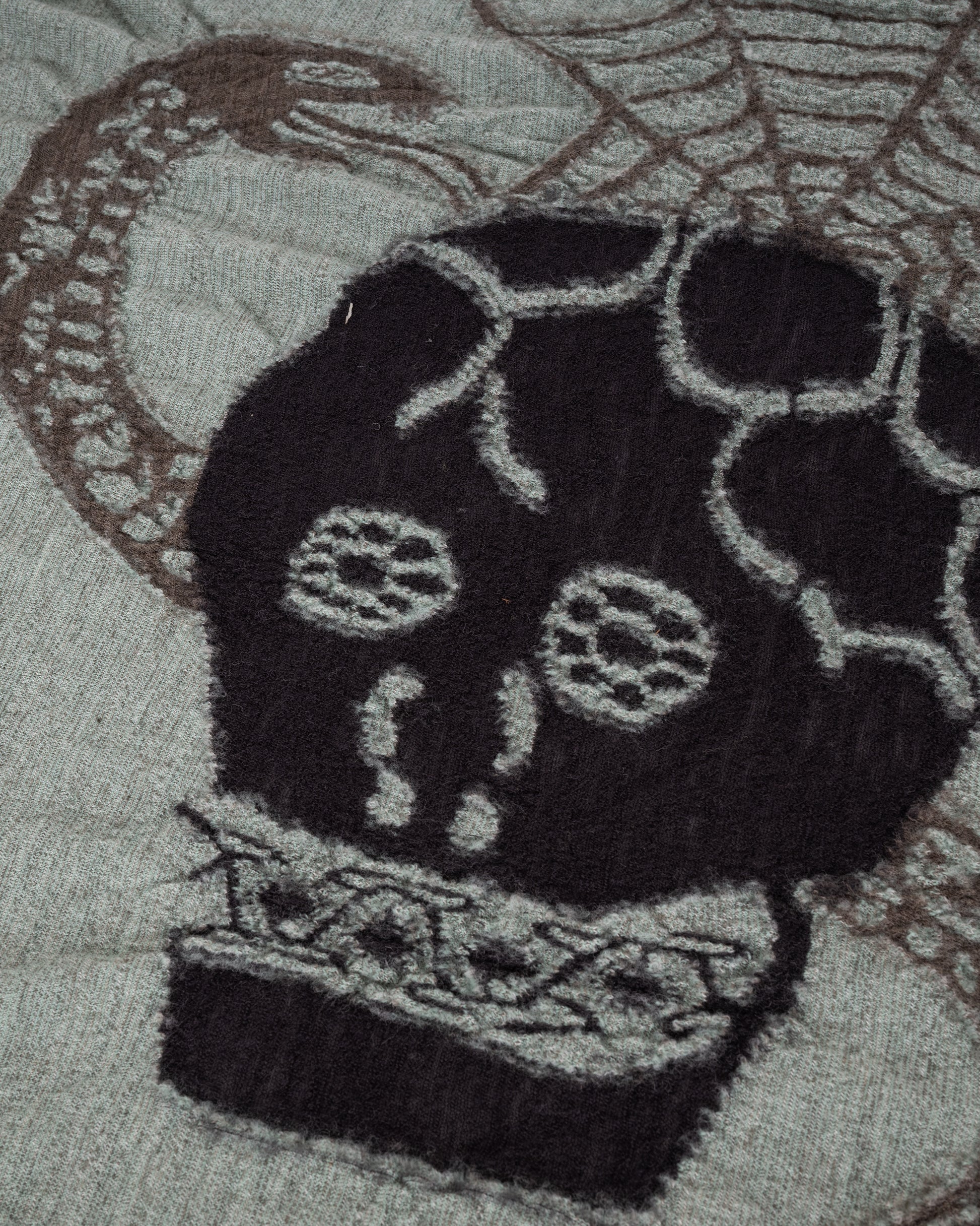 A grey and green wool scarf with a black skull in the center. Close up of the skull.