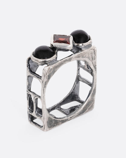 A silver square cage ring with two onyx cabochons bookending a princess cut garnet, shown from the side standing up.