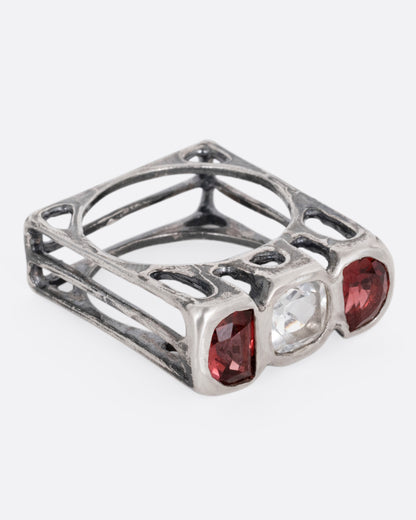 A silver ring with a cushion cut white topaz between two half moon garnets, shown from the side.