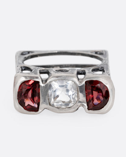 A silver ring with a cushion cut white topaz between two half moon garnets, shown from the front.