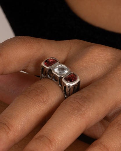 A silver ring with a cushion cut white topaz between two half moon garnets, shown on a hand.