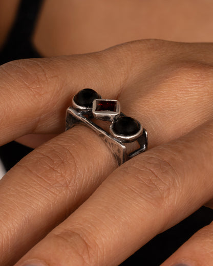 A silver square cage ring with two onyx cabochons bookending a princess cut garnet, shown on a hand.