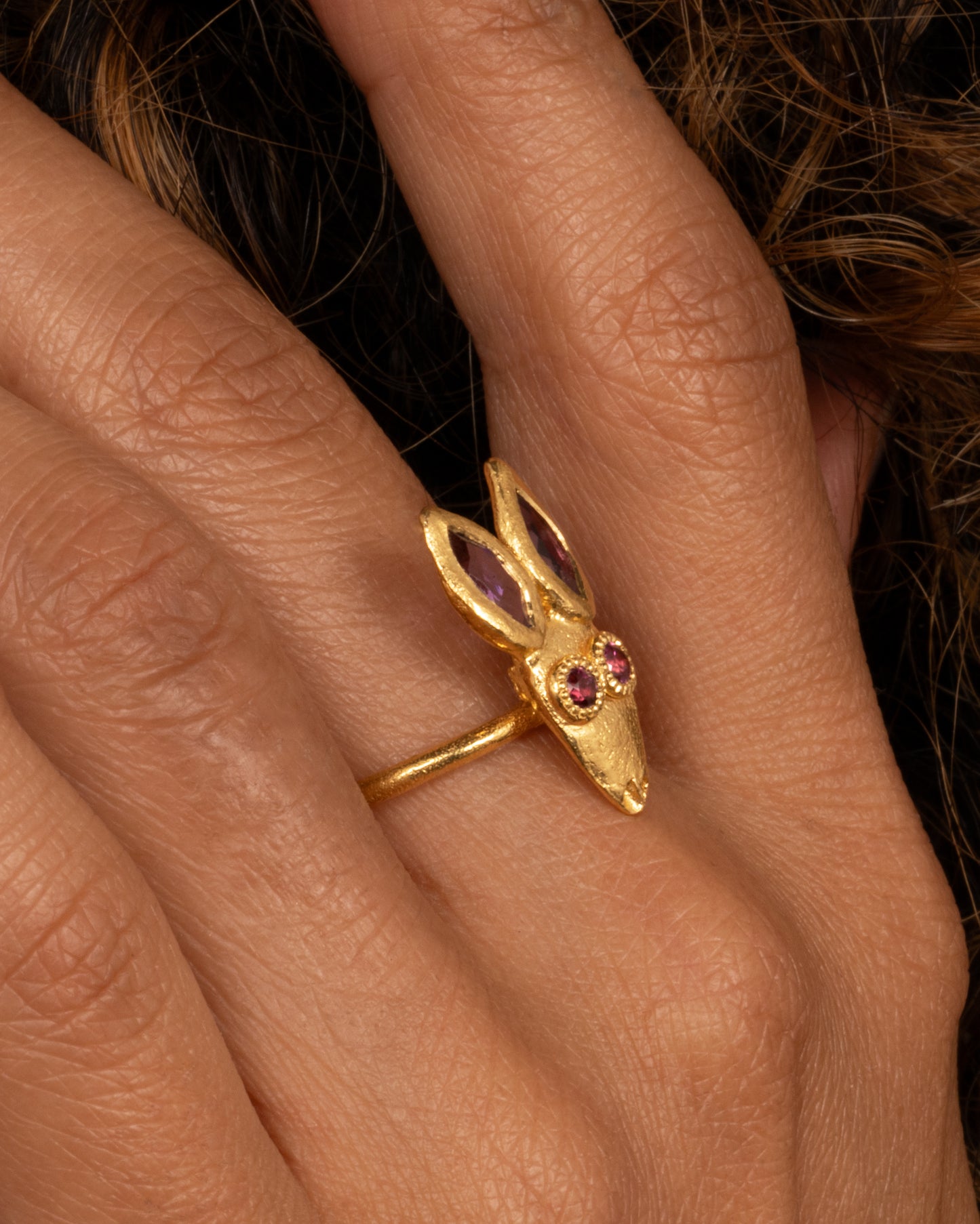 A yellow gold ring with a rabbit face with amethyst ears and tourmaline eyes. Shown on a hand.