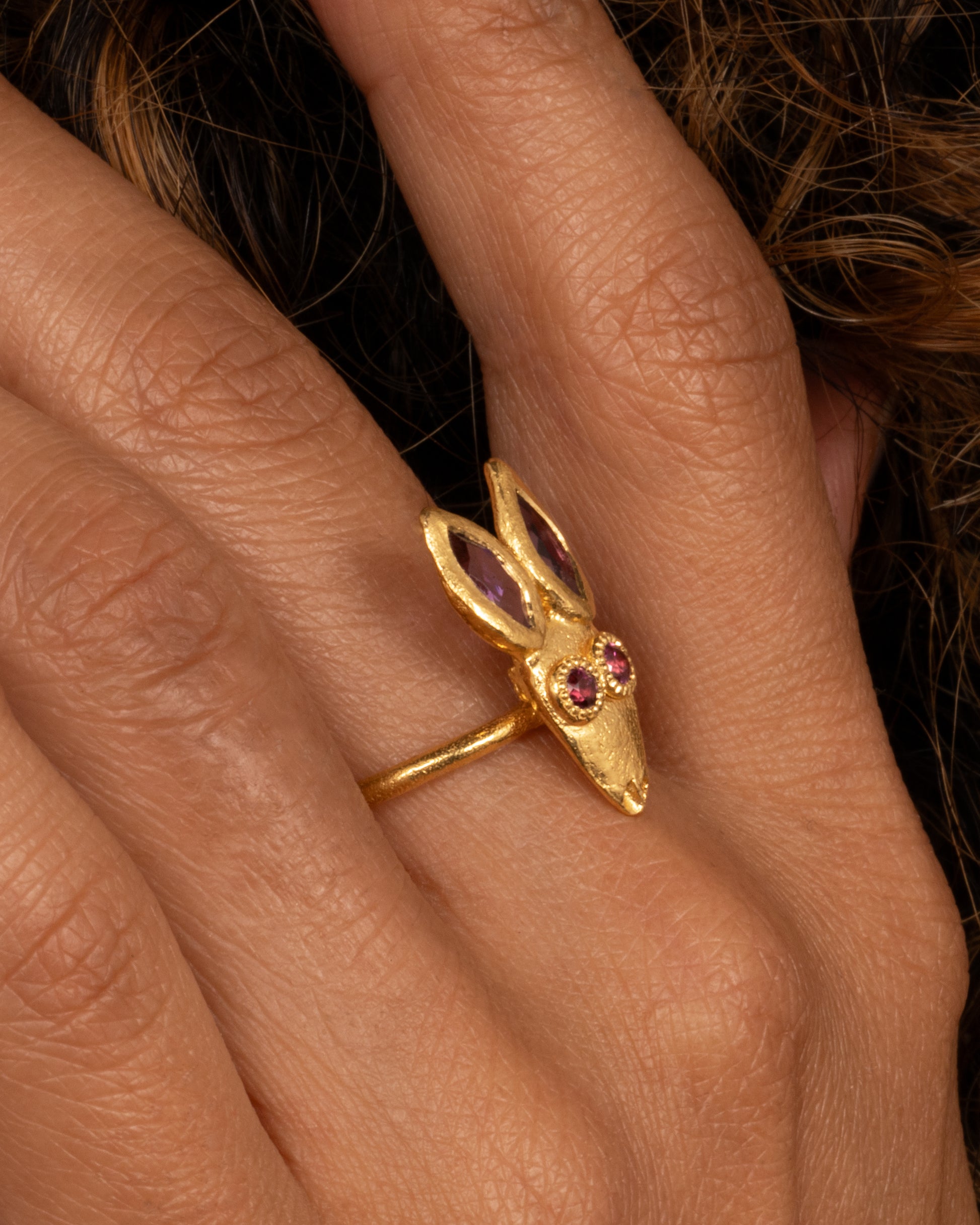 A yellow gold ring with a rabbit face with amethyst ears and tourmaline eyes. Shown on a hand.