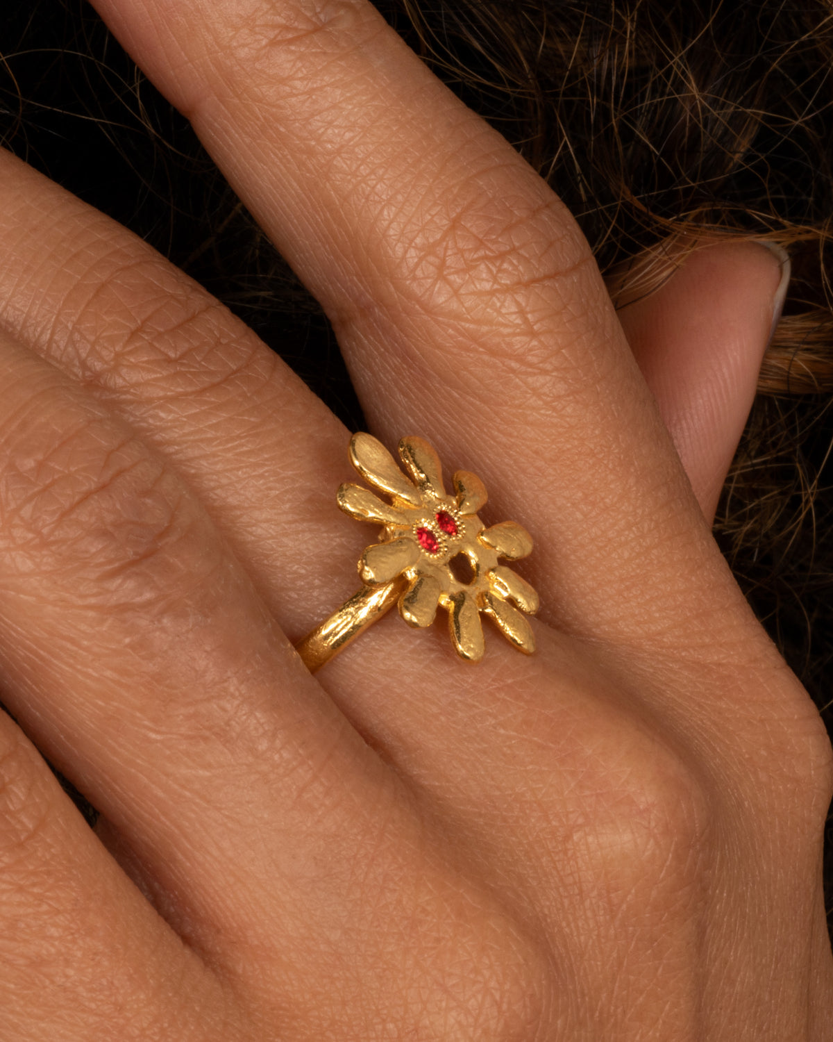 A yellow gold sun shaped ring with red stone eyes, shown on a hand.